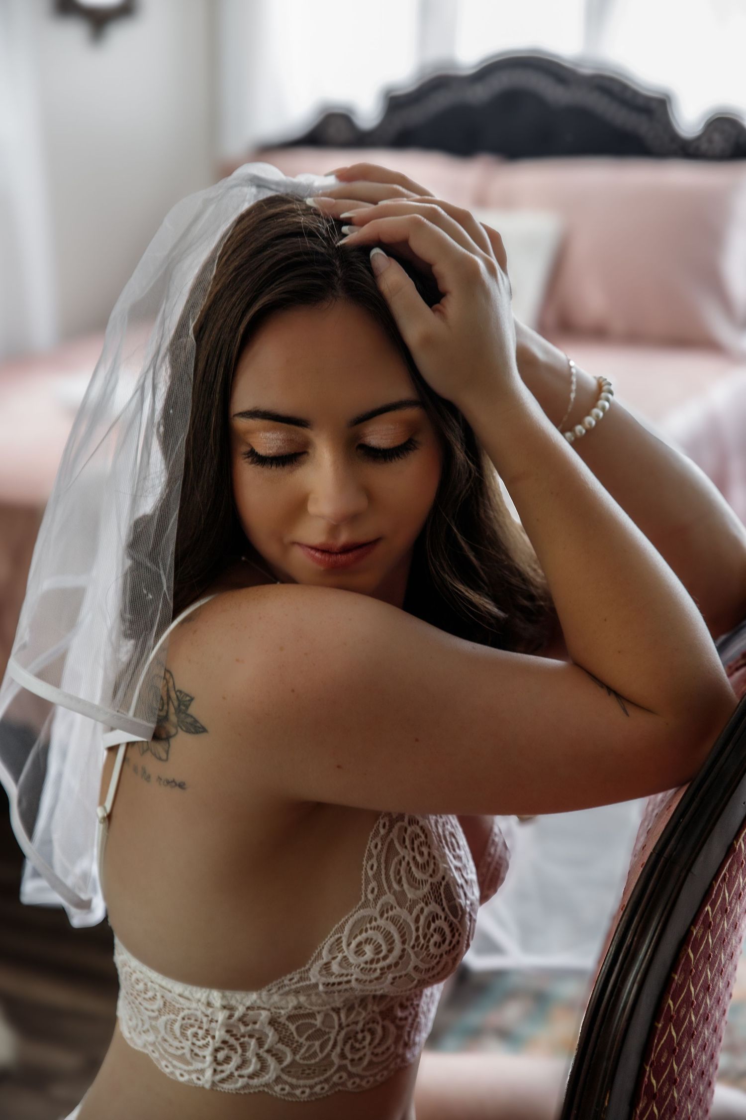 A bride in soft lighting poses in a white lace dress while touching her hair in an intimate boudoir photoshoot.
