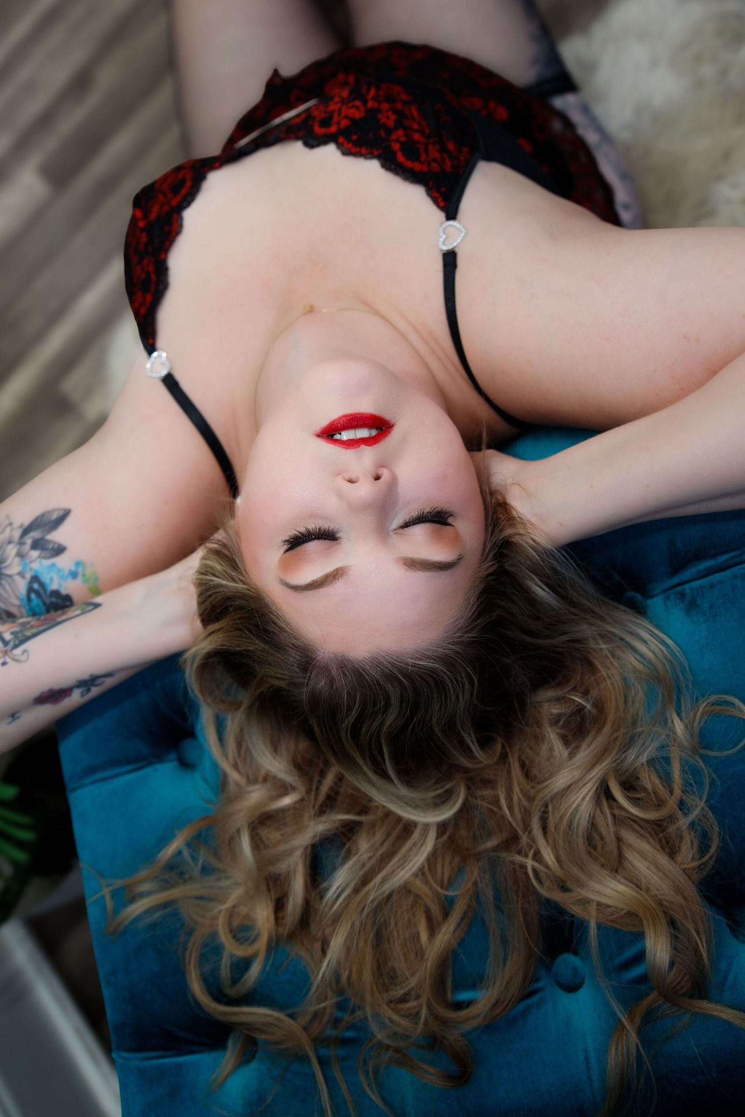 An artistic boudoir portrait showing wavy blonde hair fanned out against blue velvet with red lipstick and black lace details.