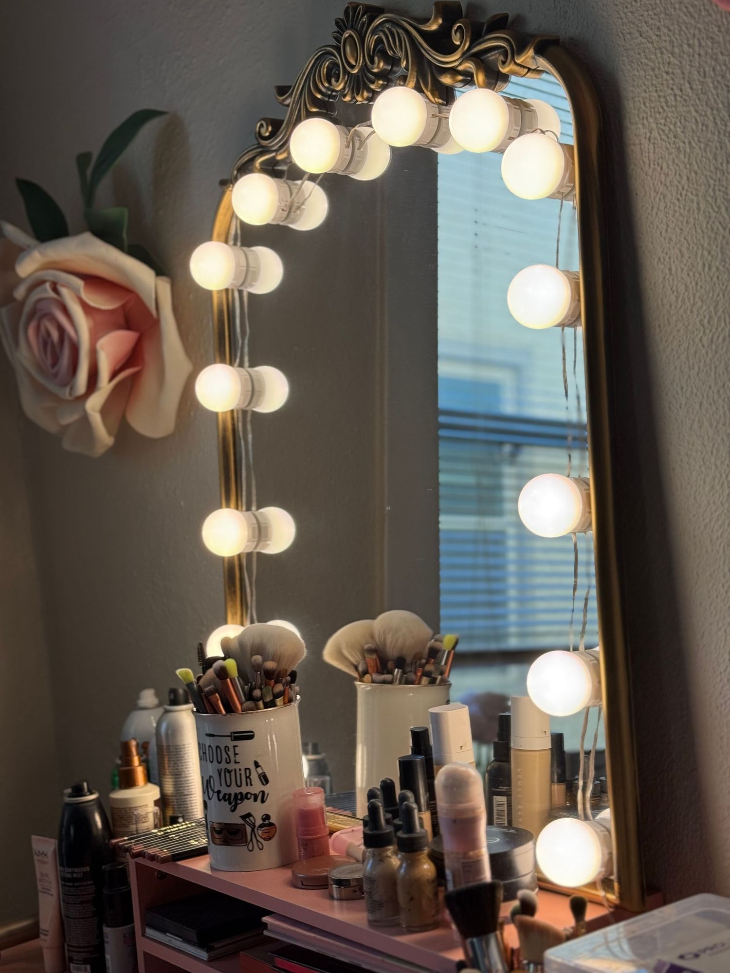 A vintage-style vanity mirror with illuminated bulbs and makeup products arranged on a wooden surface with a pink rose accent.