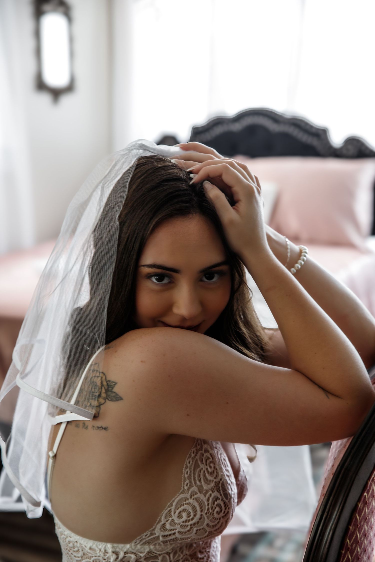 A person in a white lace wedding dress and veil with a visible shoulder tattoo poses in an intimate boudoir setting.