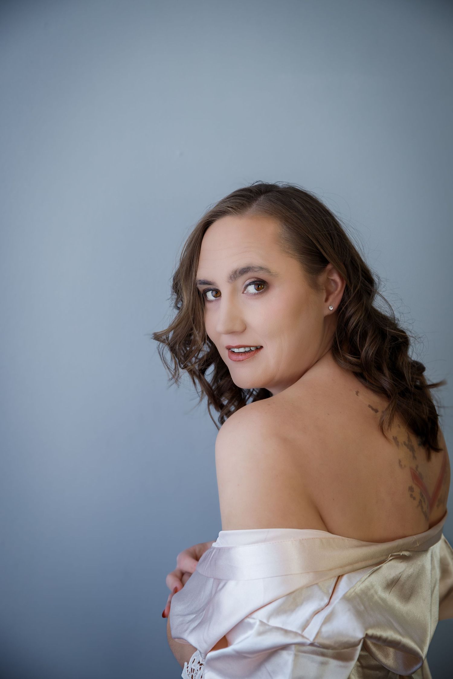 Woman in off-shoulder silk garment poses against blue background in elegant boudoir portrait with soft lighting.