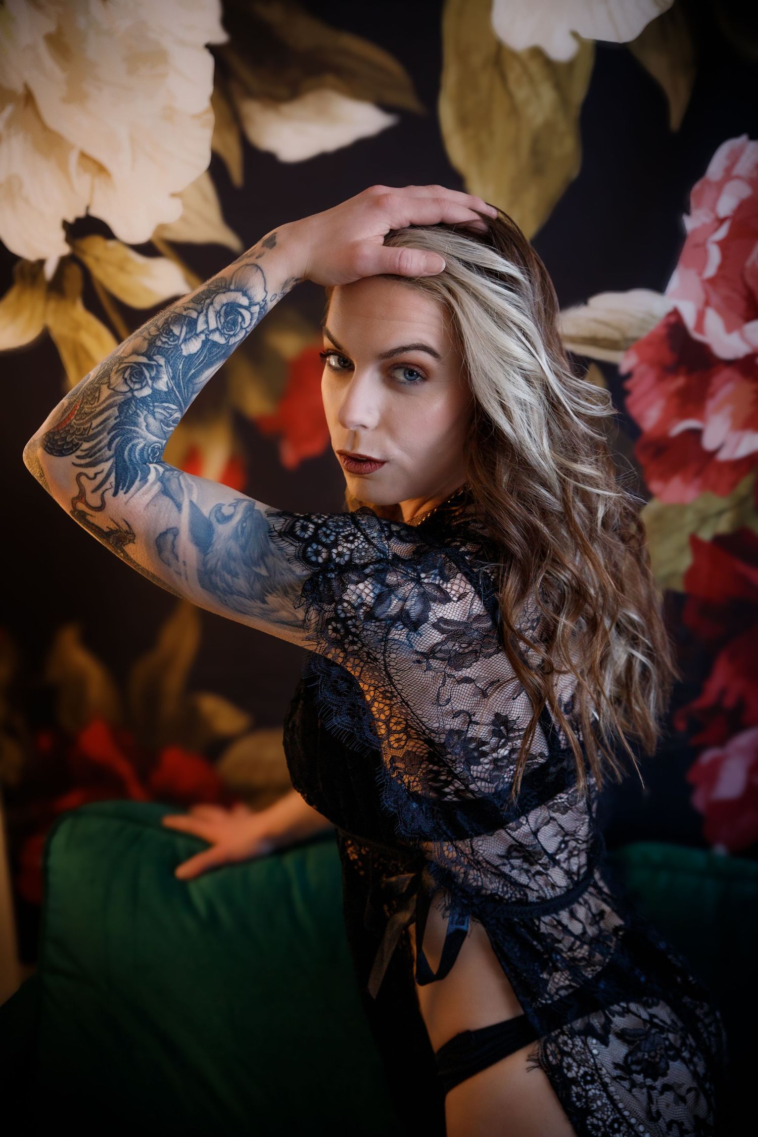Model in black lace lingerie poses dramatically against floral backdrop with visible arm tattoos.