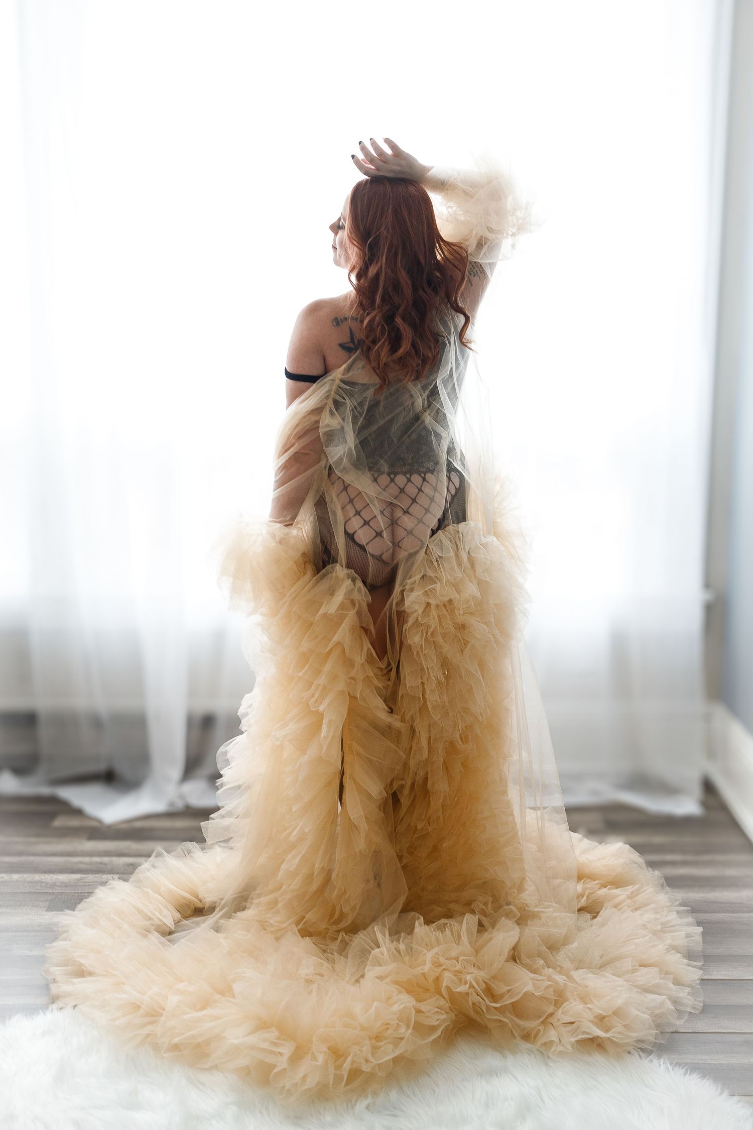 Elegant figure in flowing champagne tulle gown poses dramatically against white curtains in romantic boudoir setting.