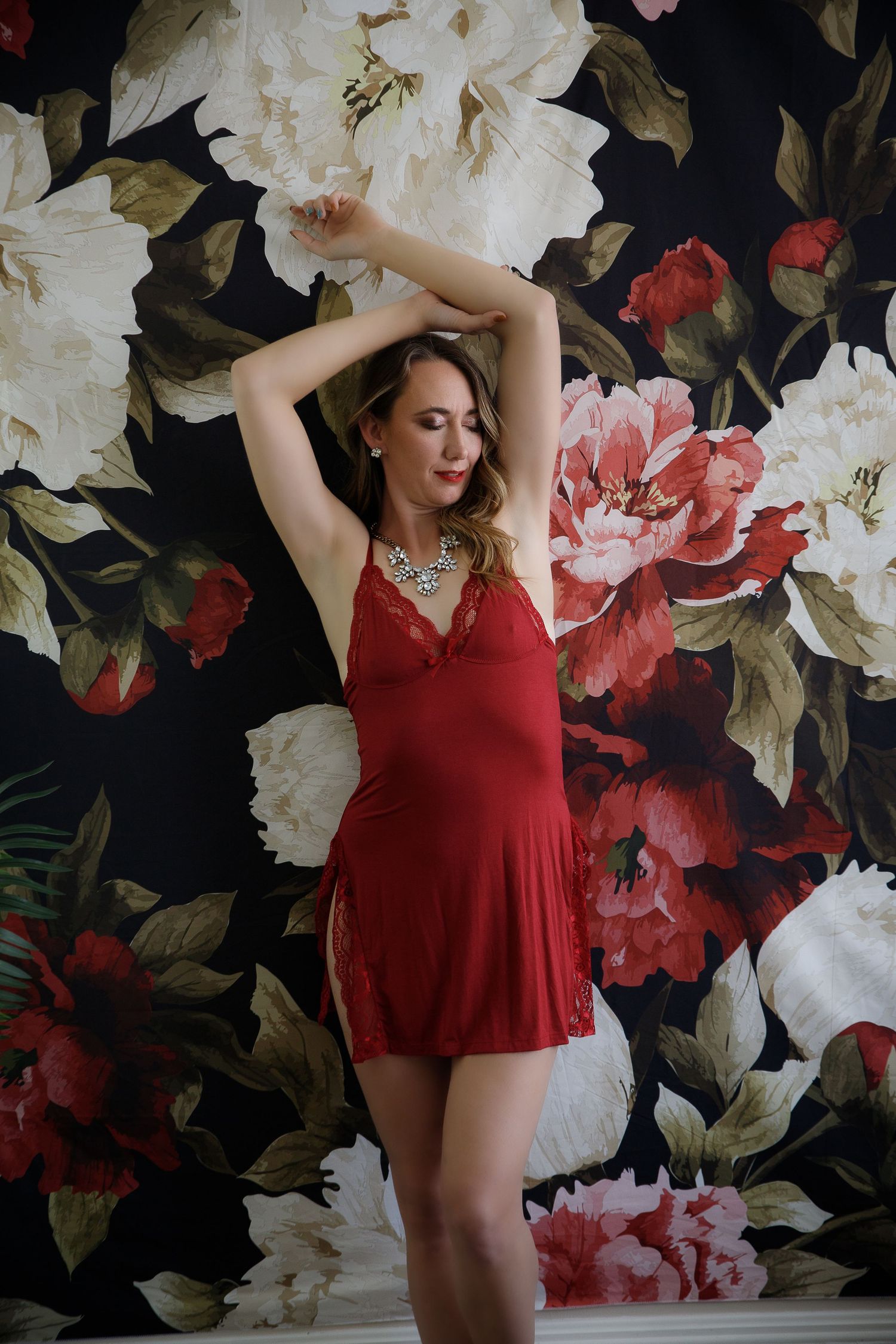 A woman in a red dress poses against a dramatic floral wallpaper with pink and white blooms on a dark background.