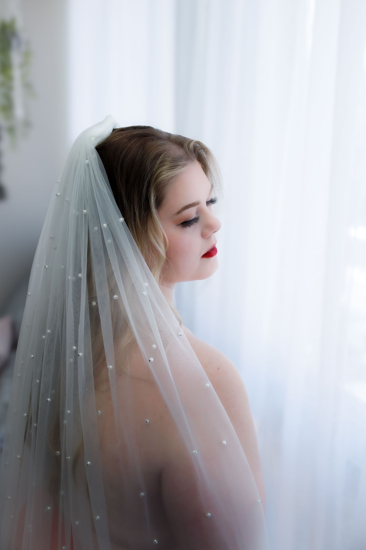 An elegant bride wearing a pearl-adorned veil looks down pensively by a bright window in soft natural light.