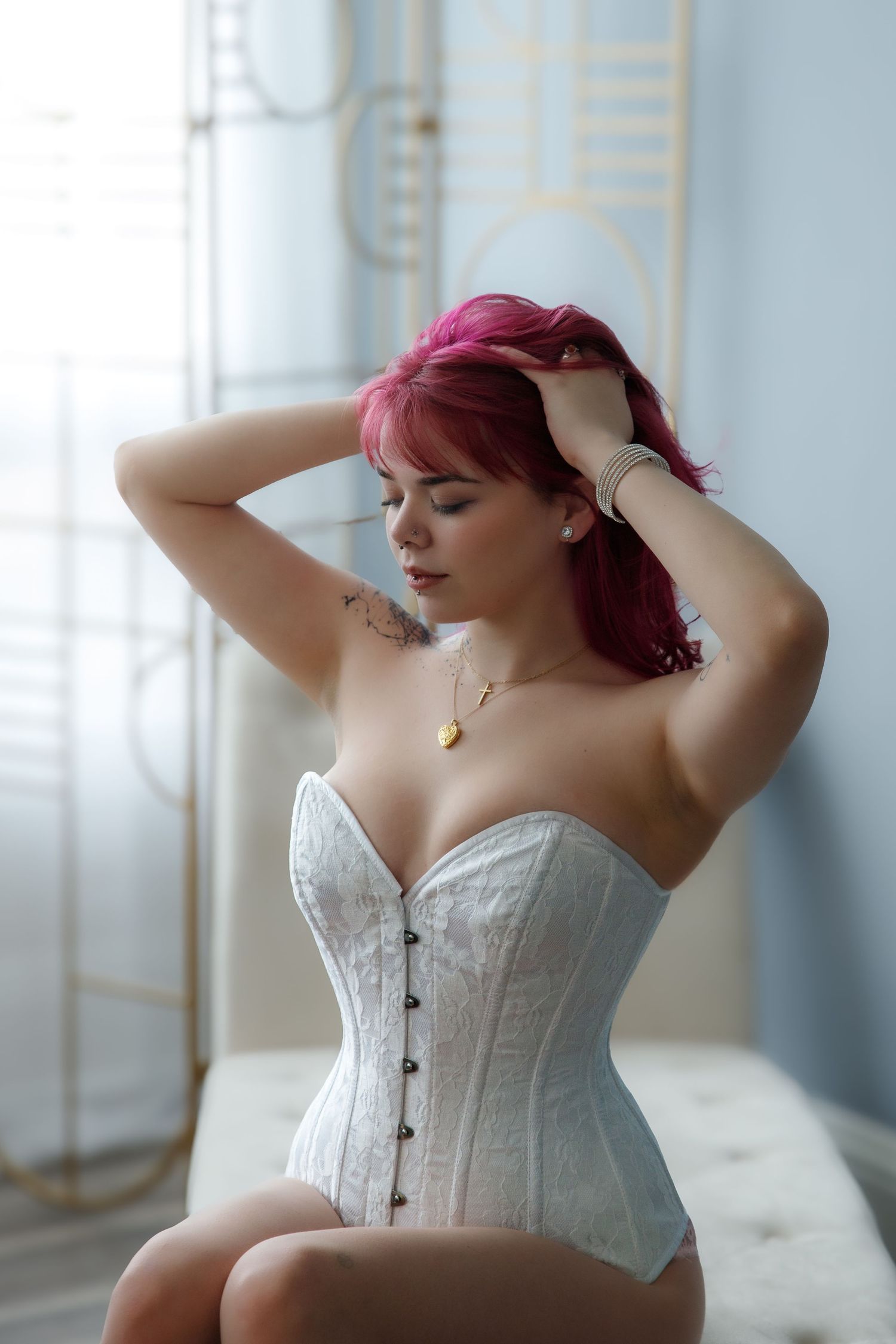 Woman with vibrant red hair poses in white lace corset near window with soft lighting.