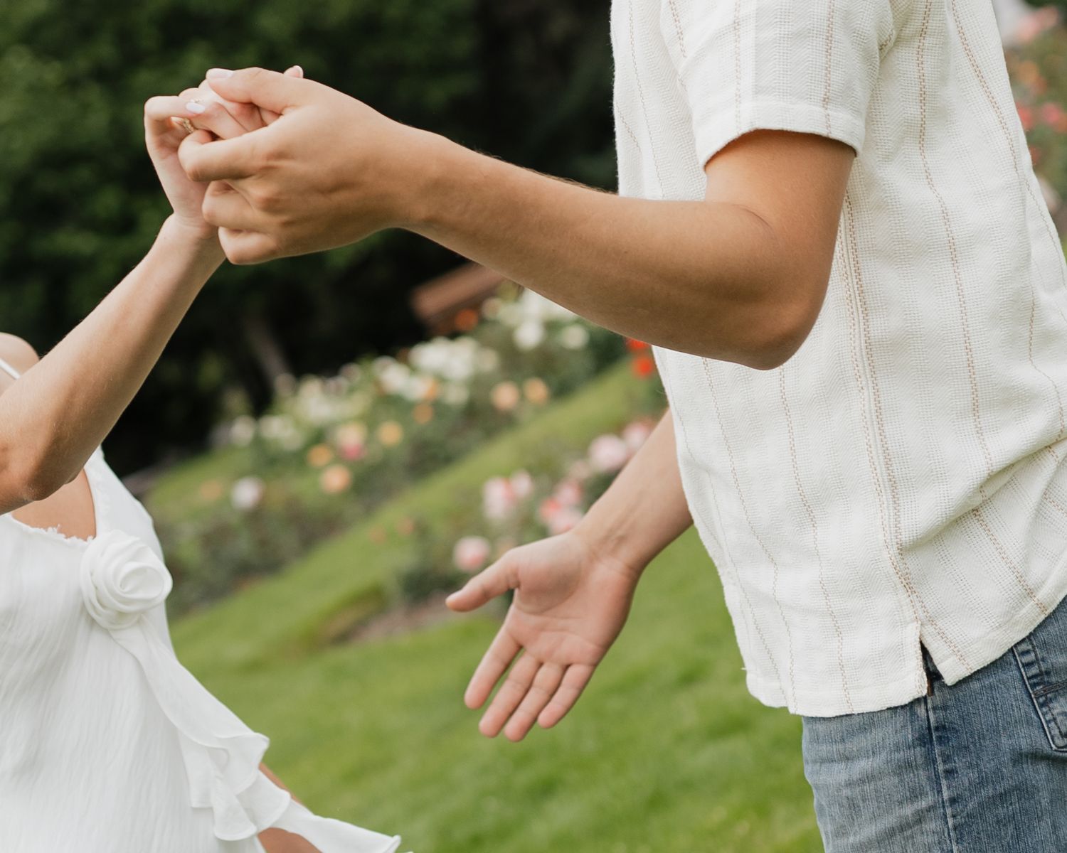 Two people hold hands outdoors in a garden with blooming flowers in the background.