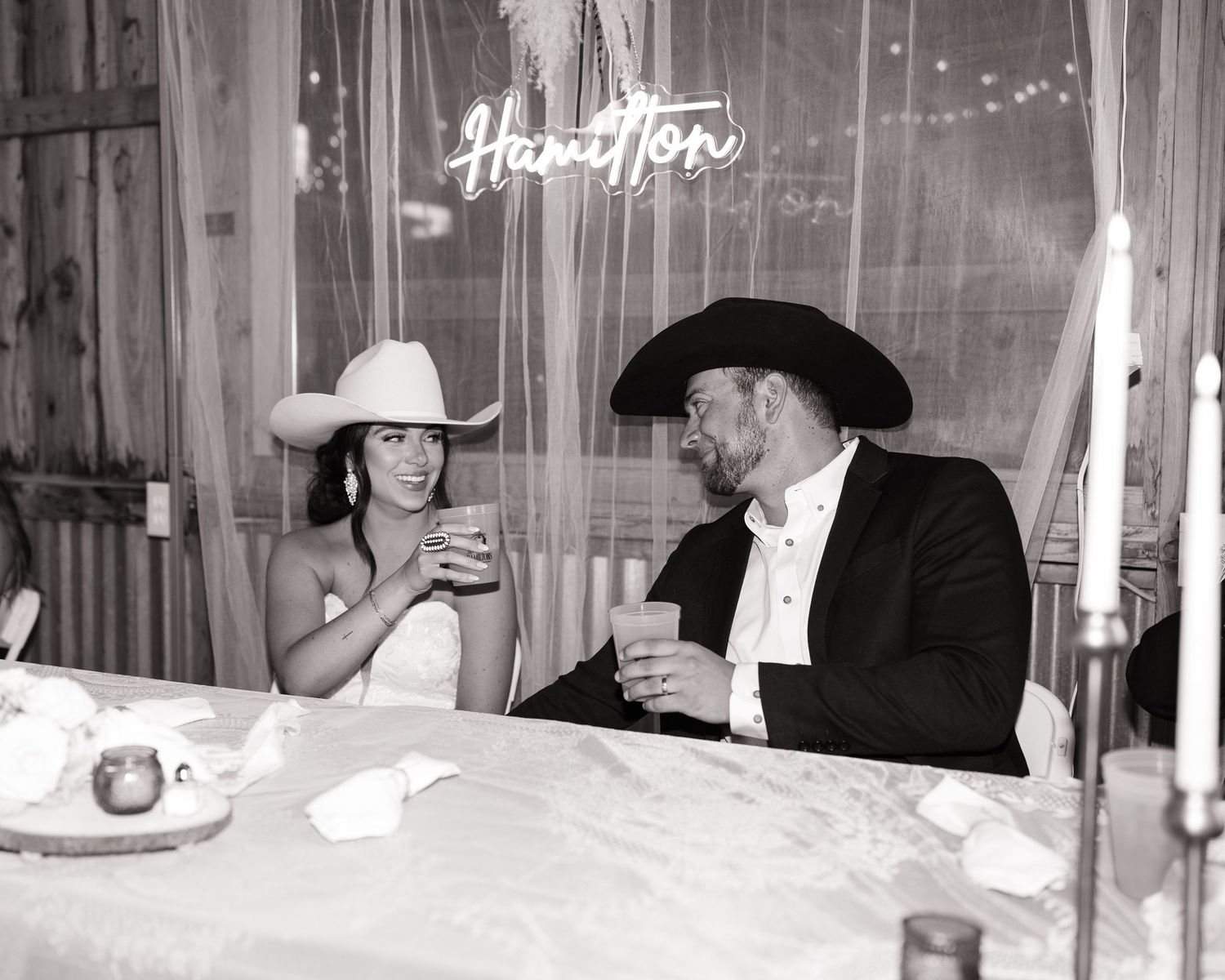 Two people in cowboy hats share drinks at a table in a black and white retro photograph.