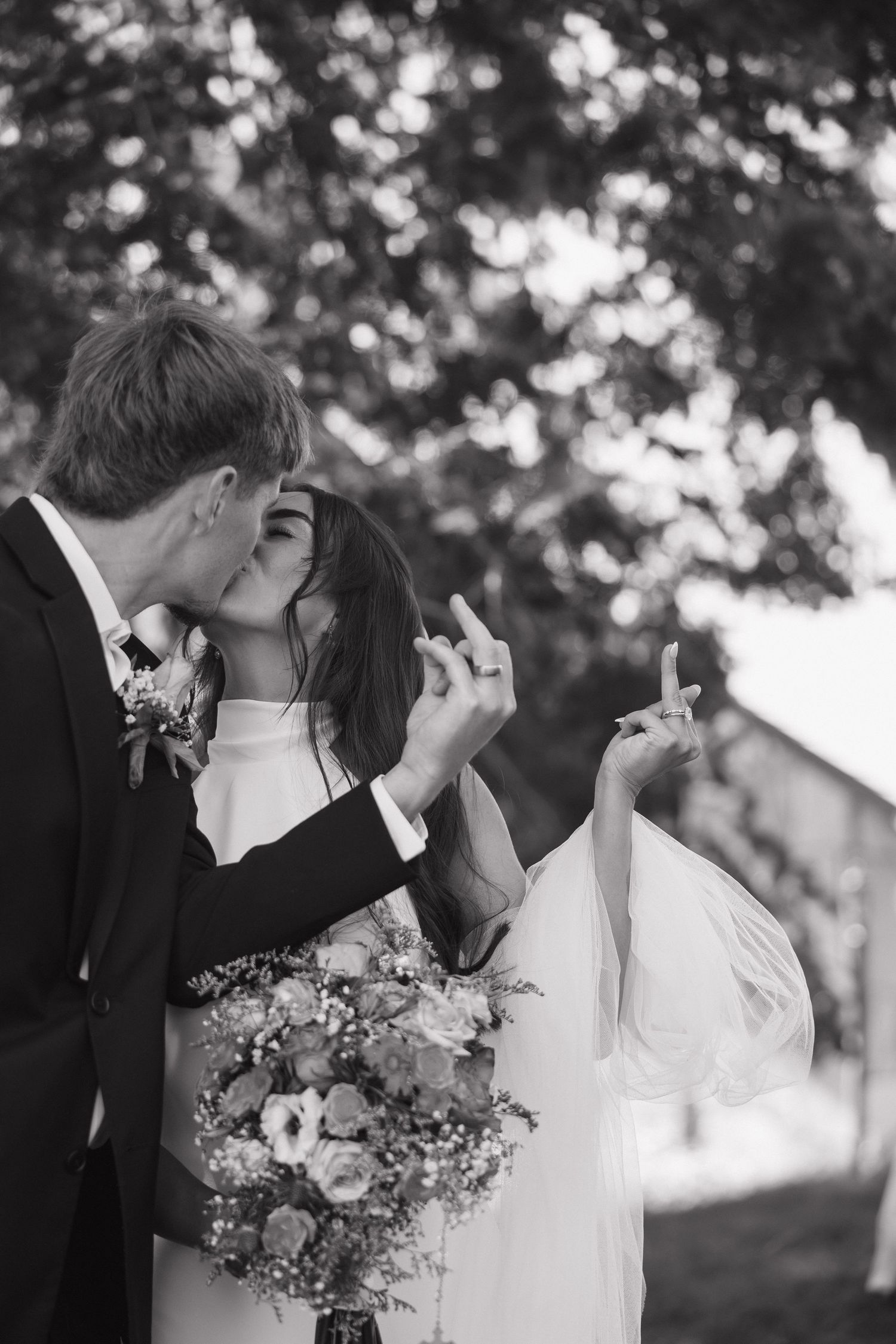 A black and white photo of a romantic wedding couple kissing while giving middle fingers in a playful moment outdoors.