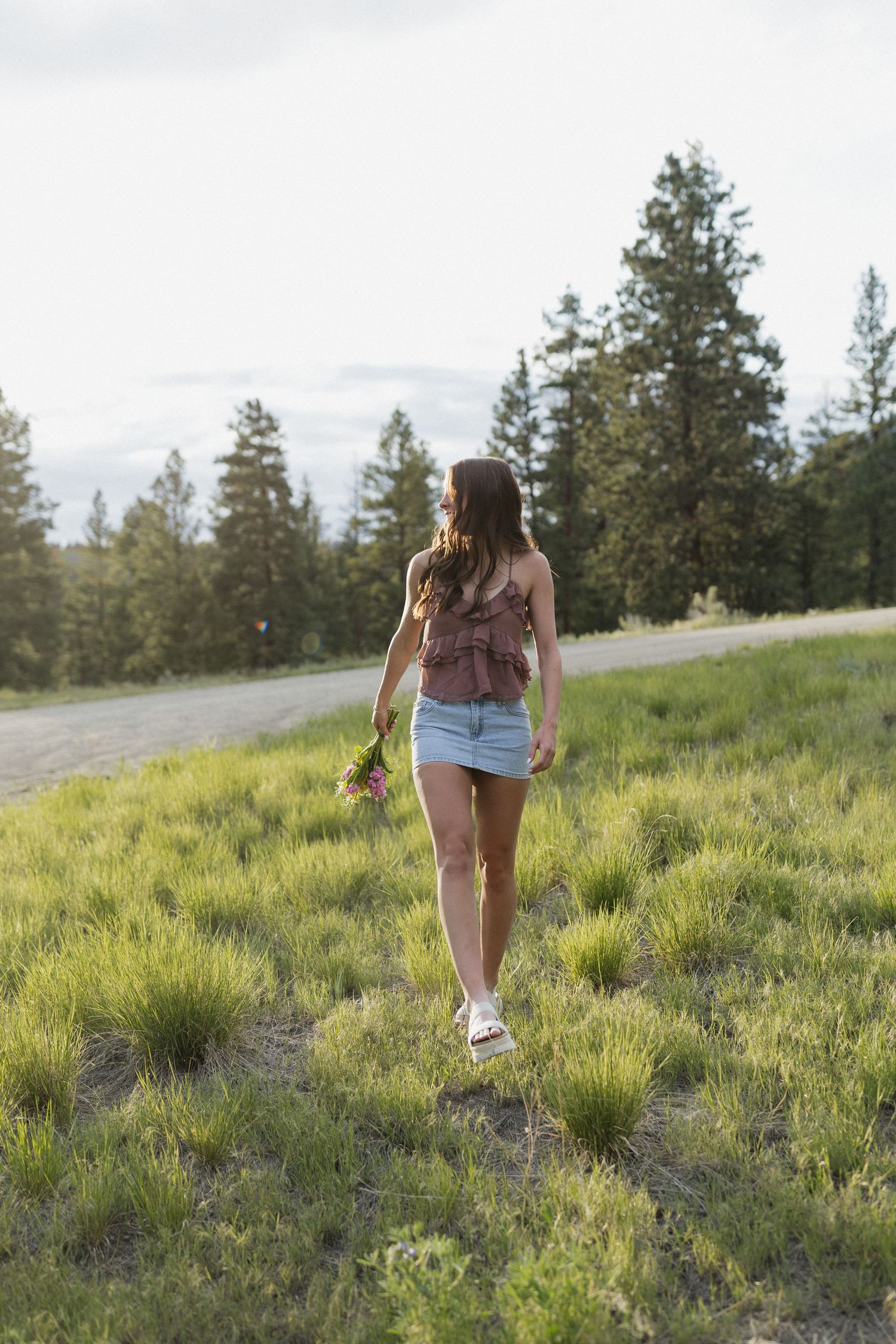 Person walking on grassy hillside holding flowers at sunset with pine trees in background.