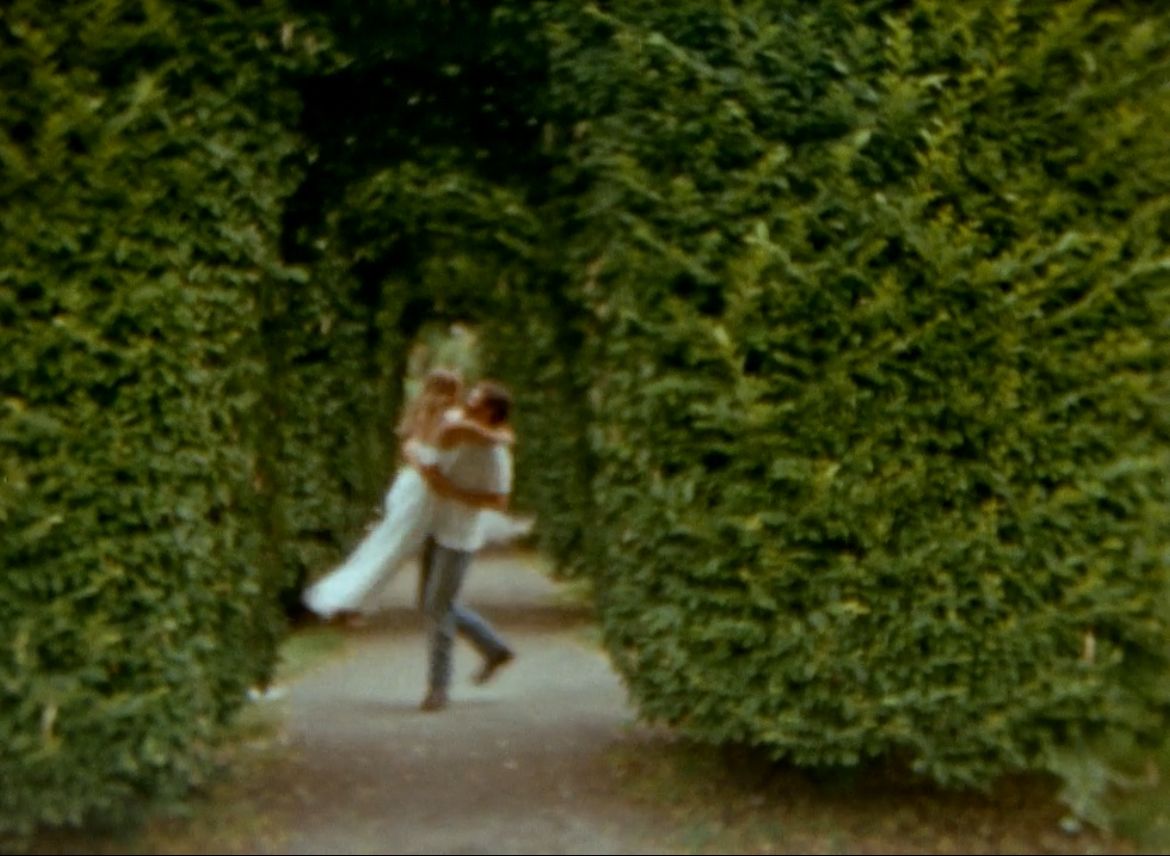 Two people dance together on a garden path surrounded by tall green hedges in a dreamy outdoor setting.