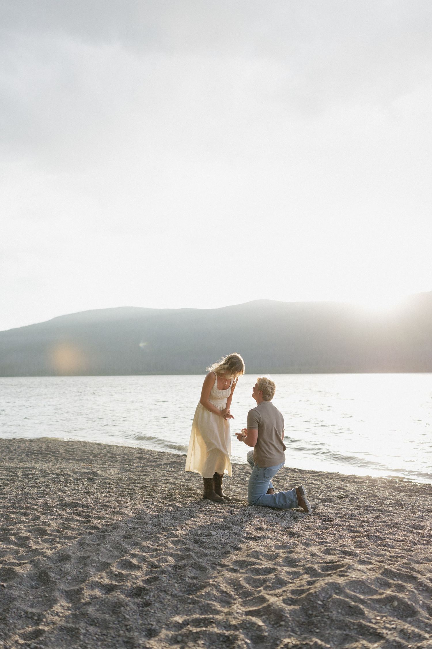 A couple shares an intimate moment during sunset on a sandy beach beside a serene lake with mountains in the background.