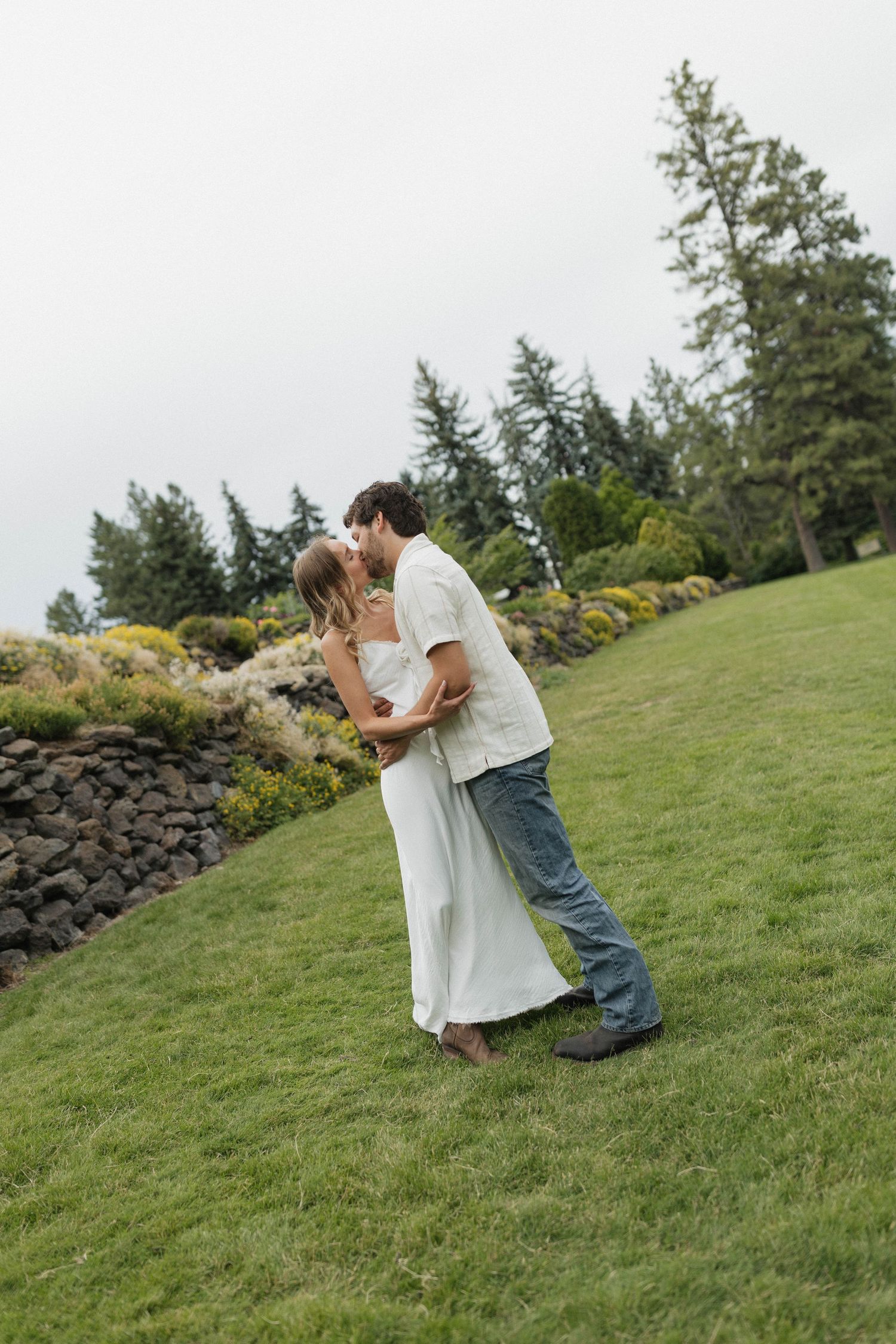 A romantic couple embrace and kiss on a grassy hillside with evergreen trees in the background.