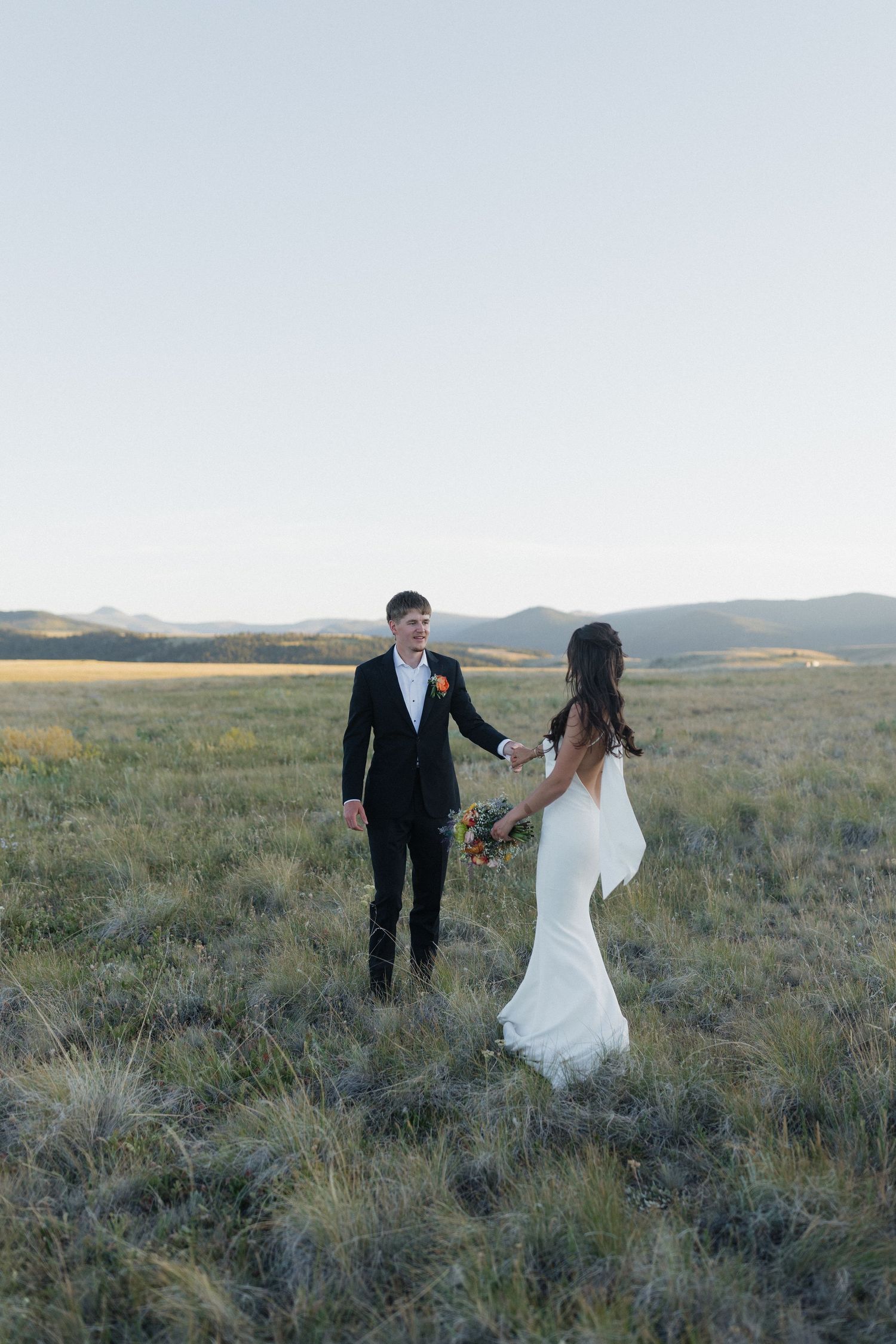 A wedding couple stands together holding hands in a scenic mountain meadow at dusk.