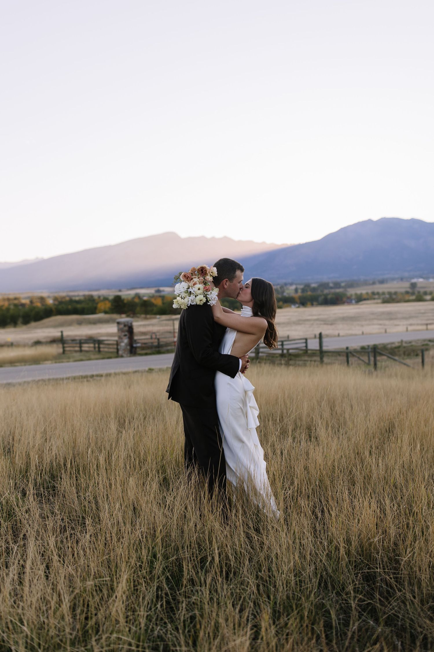 A couple embraces in a field of tall grass at sunset with mountains in the background.