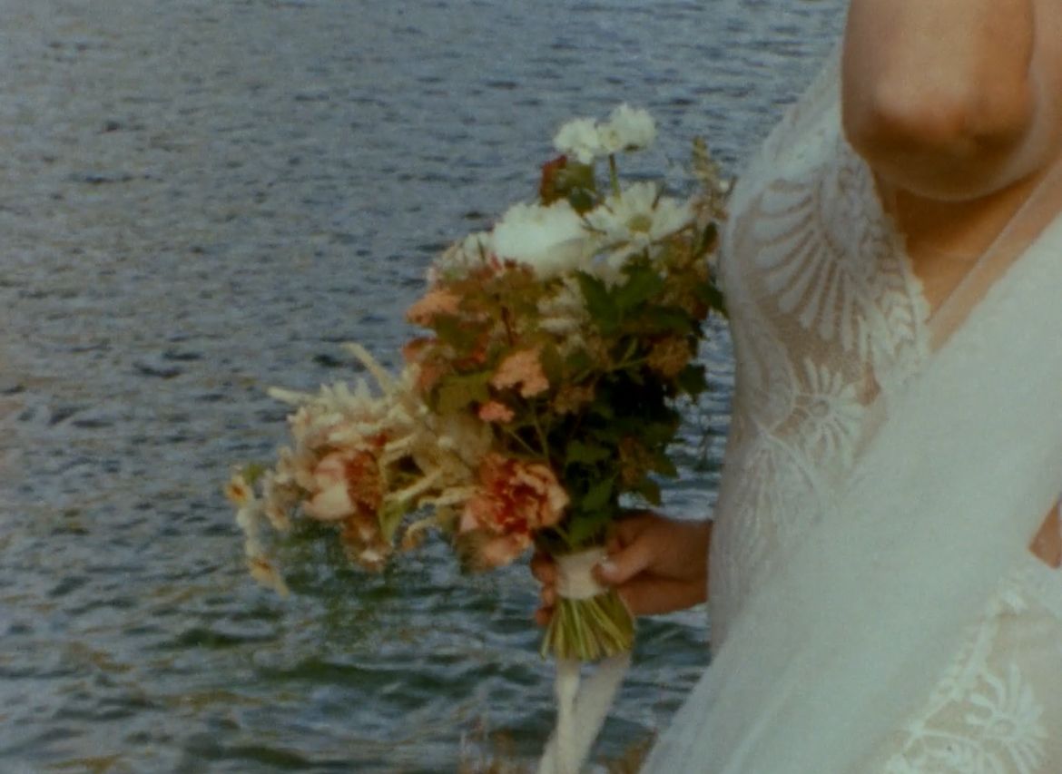 A wedding bouquet with soft peach and white flowers is held against a white lace dress near a rippling water surface.