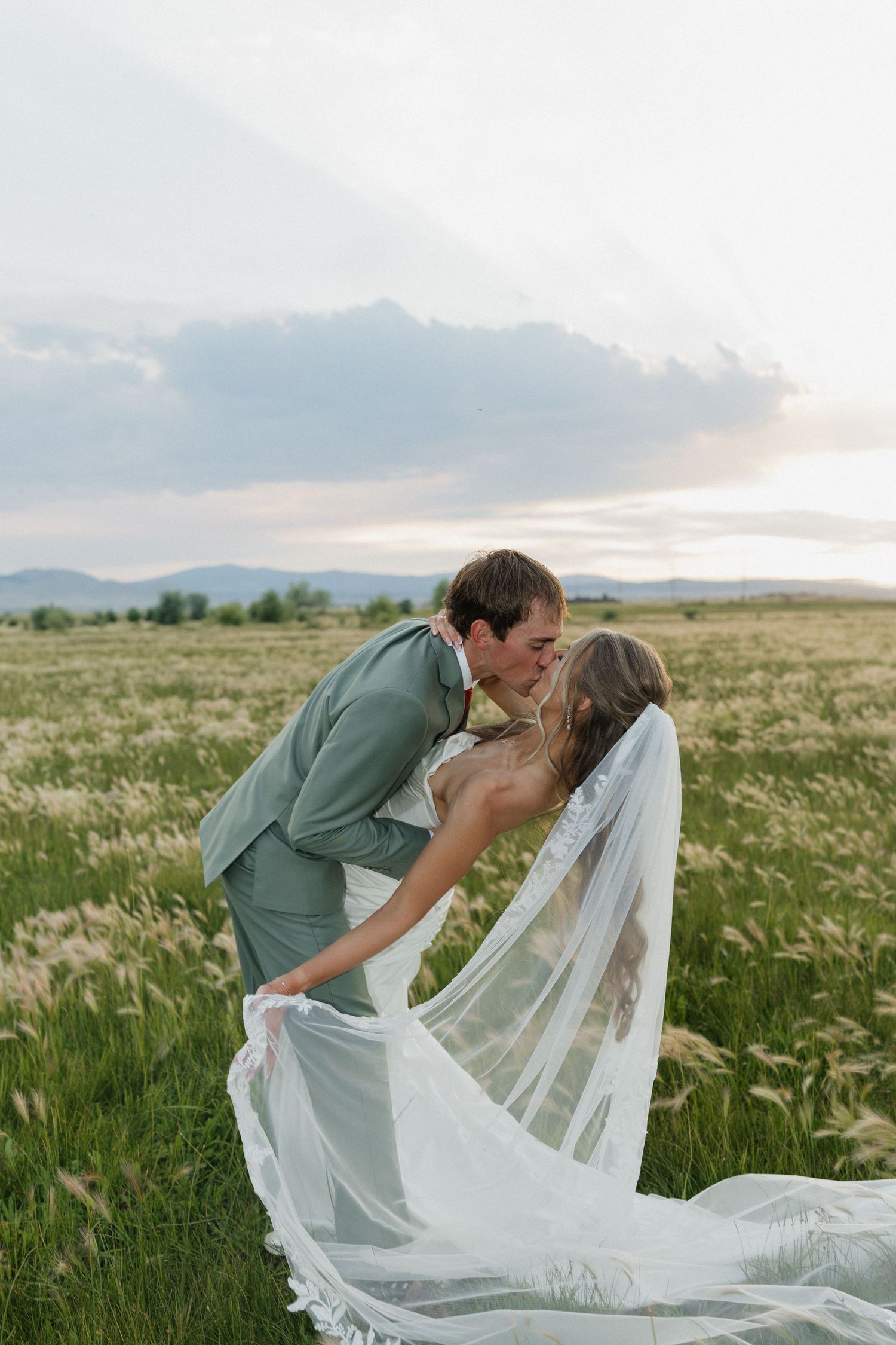 A couple shares a romantic kiss while the bride's veil flows in the wind across a scenic mountain meadow landscape.