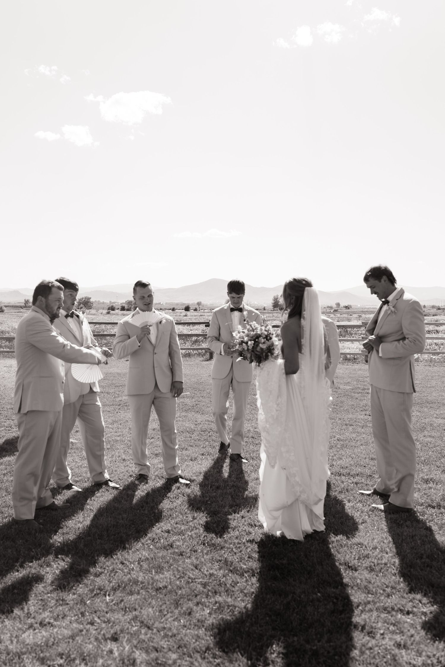A wedding party stands together in black and white on an outdoor field casting long shadows across the grass.