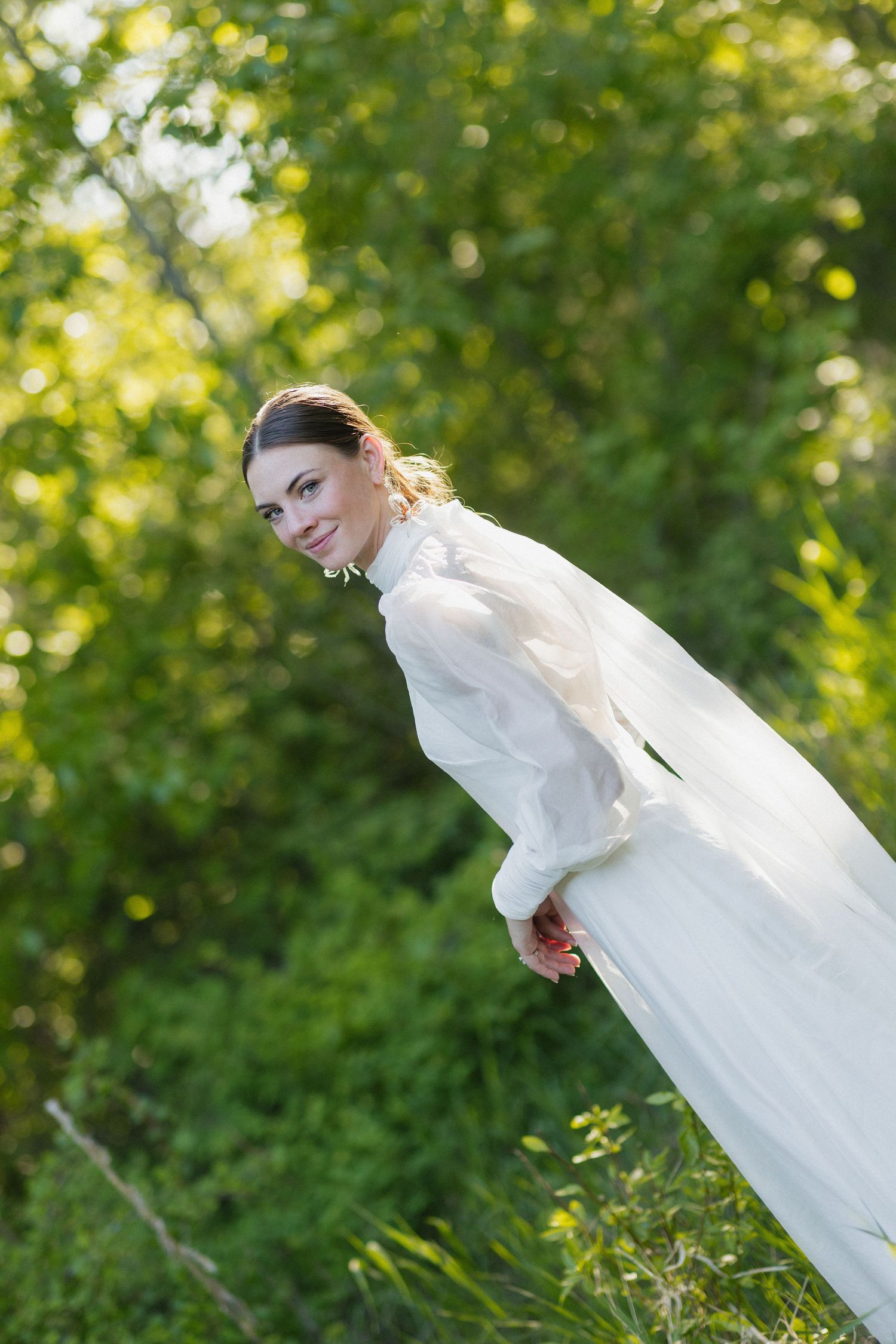 A bride in a classic white long-sleeved wedding gown poses playfully outdoors against a sunlit green background.
