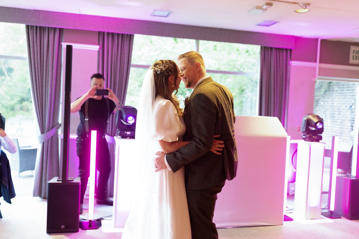 Two people share their first dance as newlyweds in a reception hall with purple ambient lighting and a DJ in background.