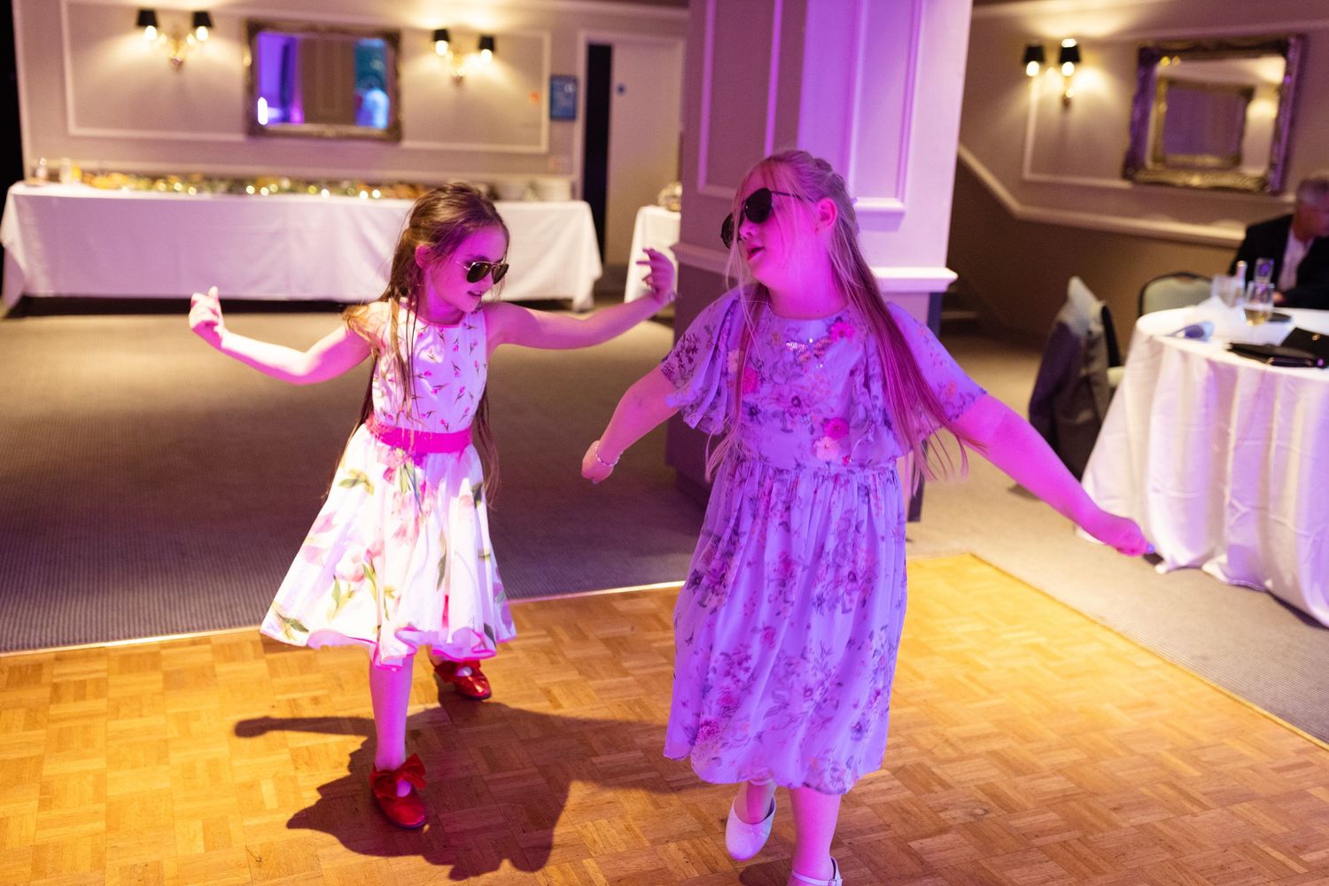 Two young dancers in floral dresses twirl together on a wooden dance floor at a formal event with purple lighting.