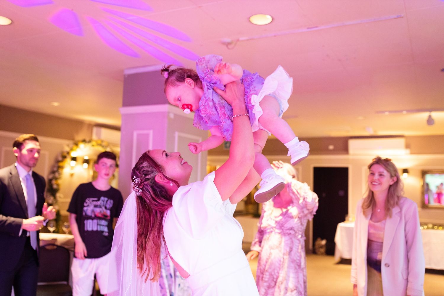 A person in purple dance attire is lifted up at an indoor dance celebration under pink and purple lighting.