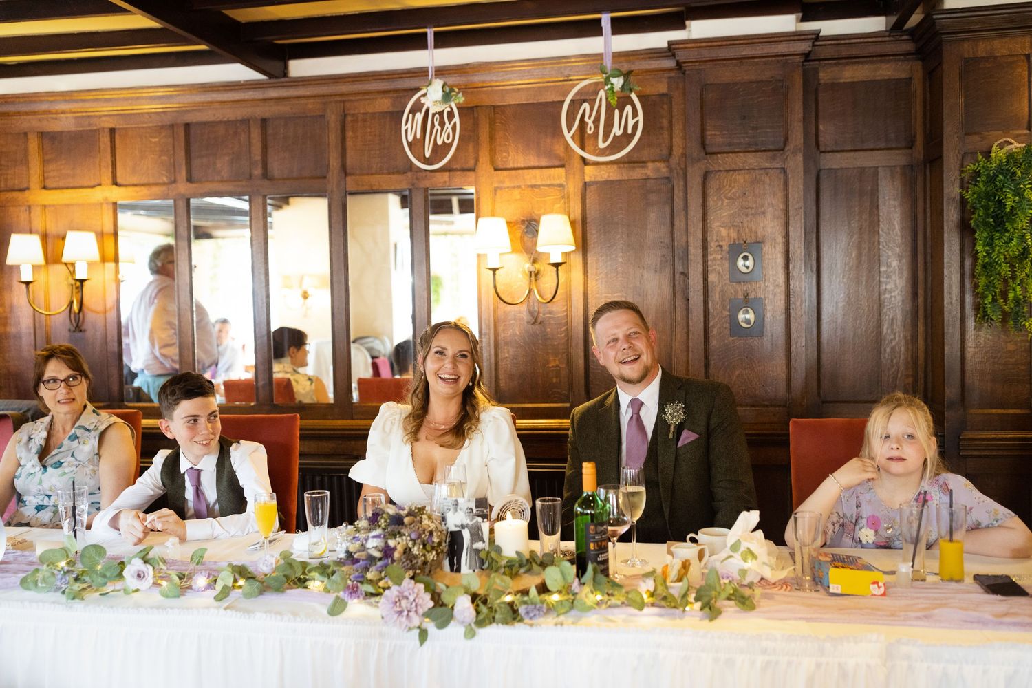 Wedding party members smile while seated at an elegant head table with floral decorations and glowing candles.