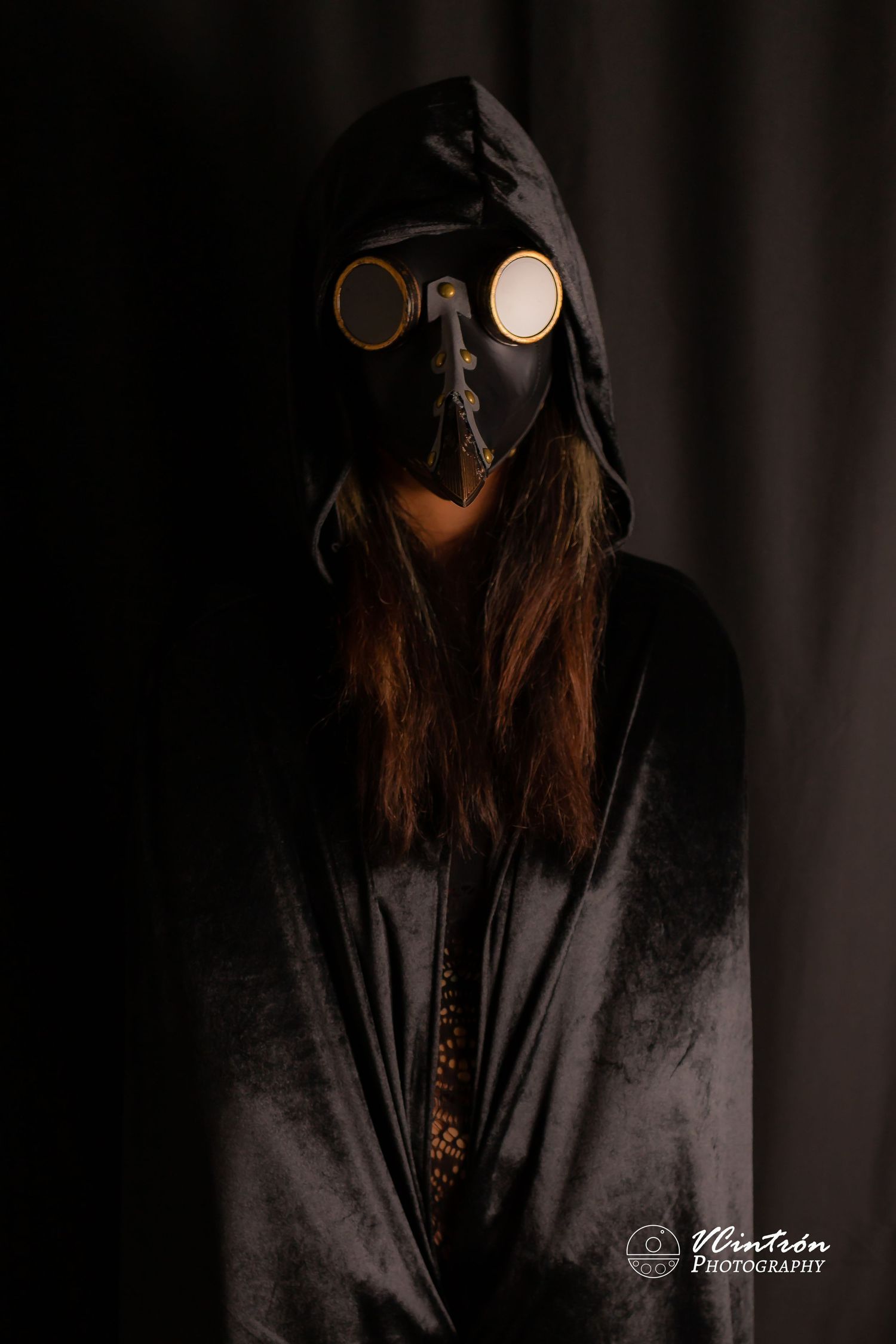 Dark moody portrait of a figure in a gas mask wearing black clothing with long hair against a shadowy background.