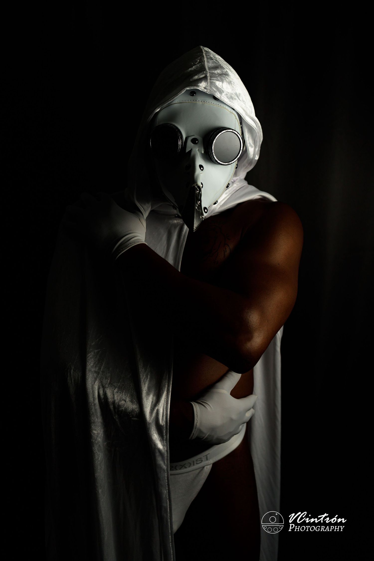 Dramatic low-key portrait of a figure wearing a white plague doctor mask and cape against a dark background.