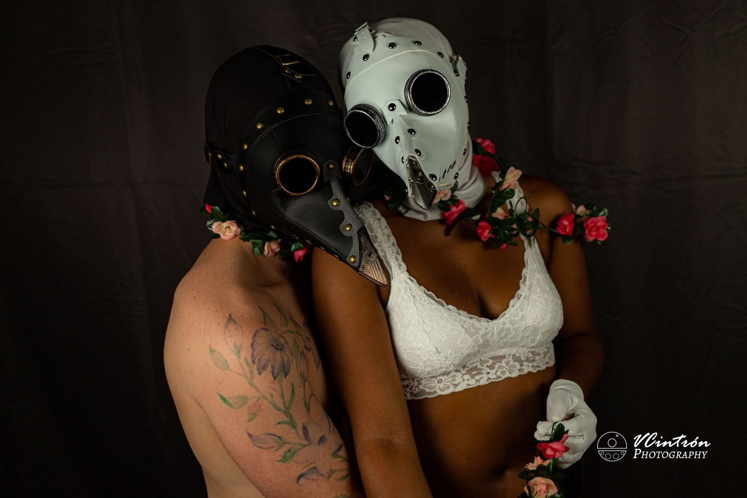 Two people in lace plague doctor masks and flowers embrace against dark background for romantic gothic photoshoot.