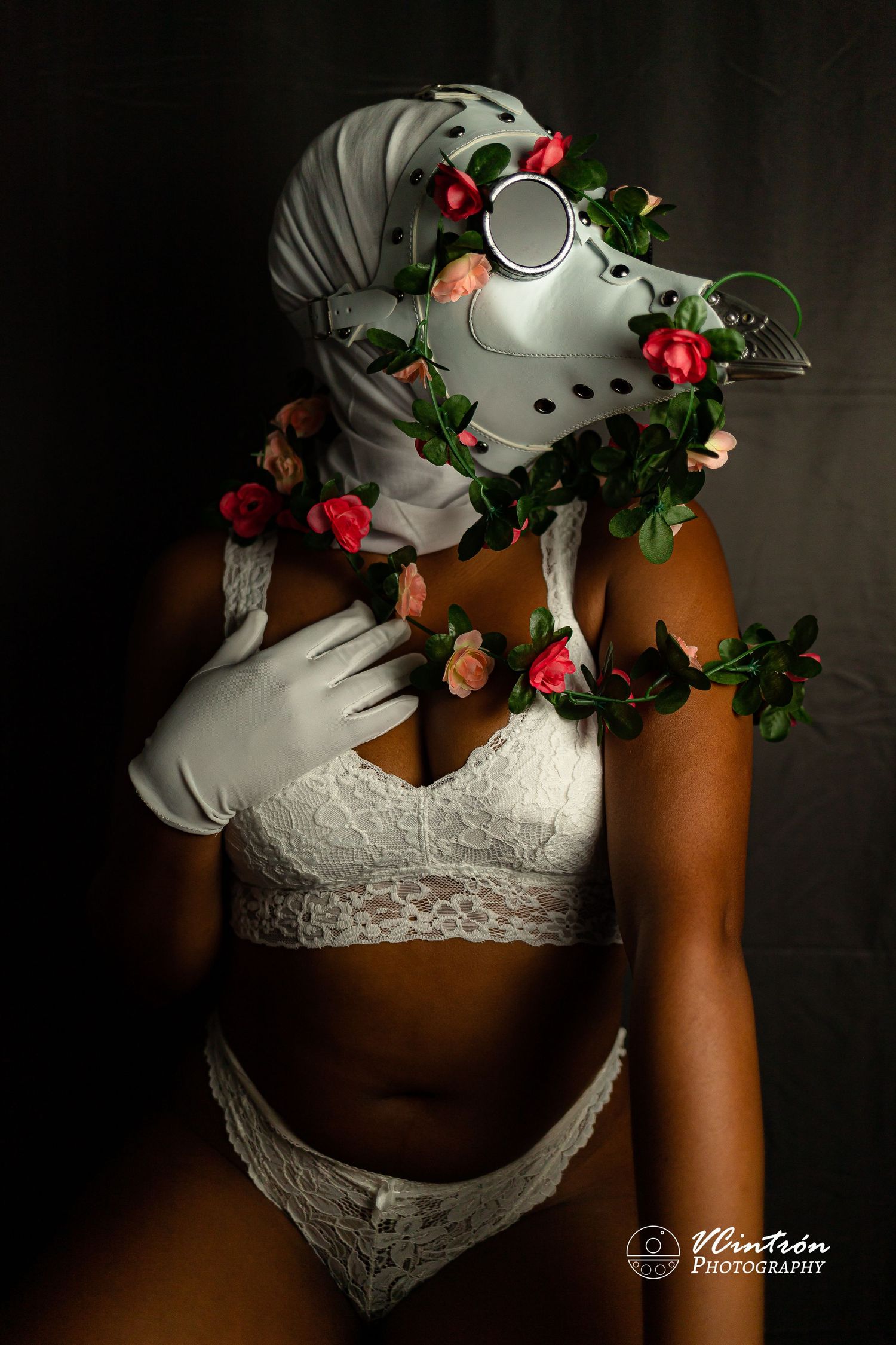 Artistic portrait featuring white lace lingerie, metallic plague doctor mask, and climbing red roses against dark background.