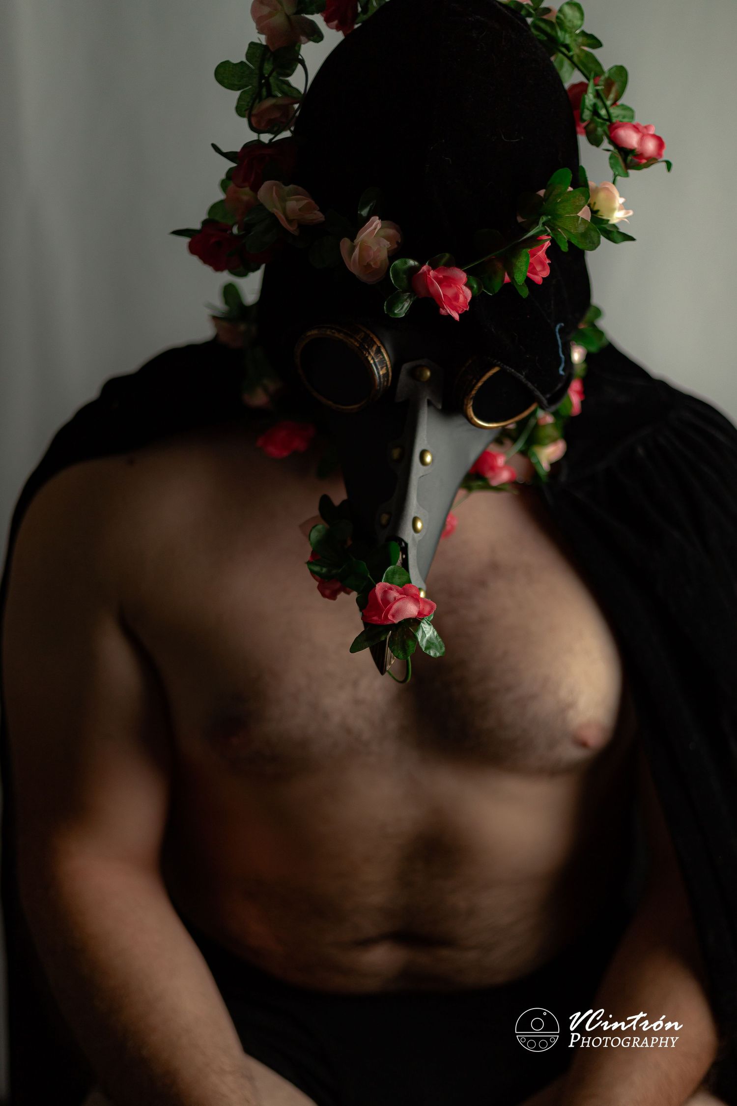 Dark, moody artistic portrait with decorative floral crown and mask adorned in deep red roses and green foliage.
