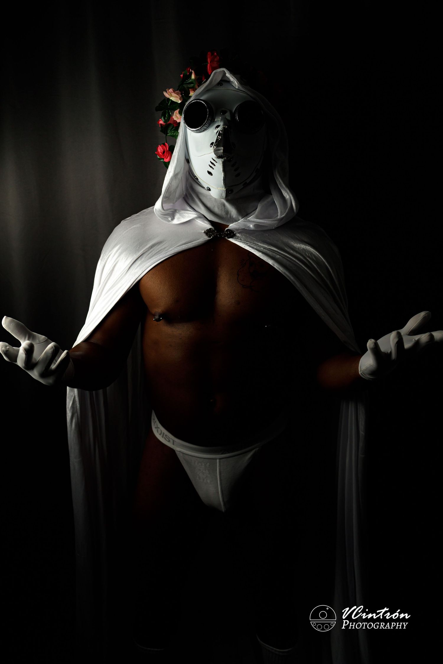 Dramatic low-key photo of a muscular figure in white robes and mask striking a powerful pose against dark background.
