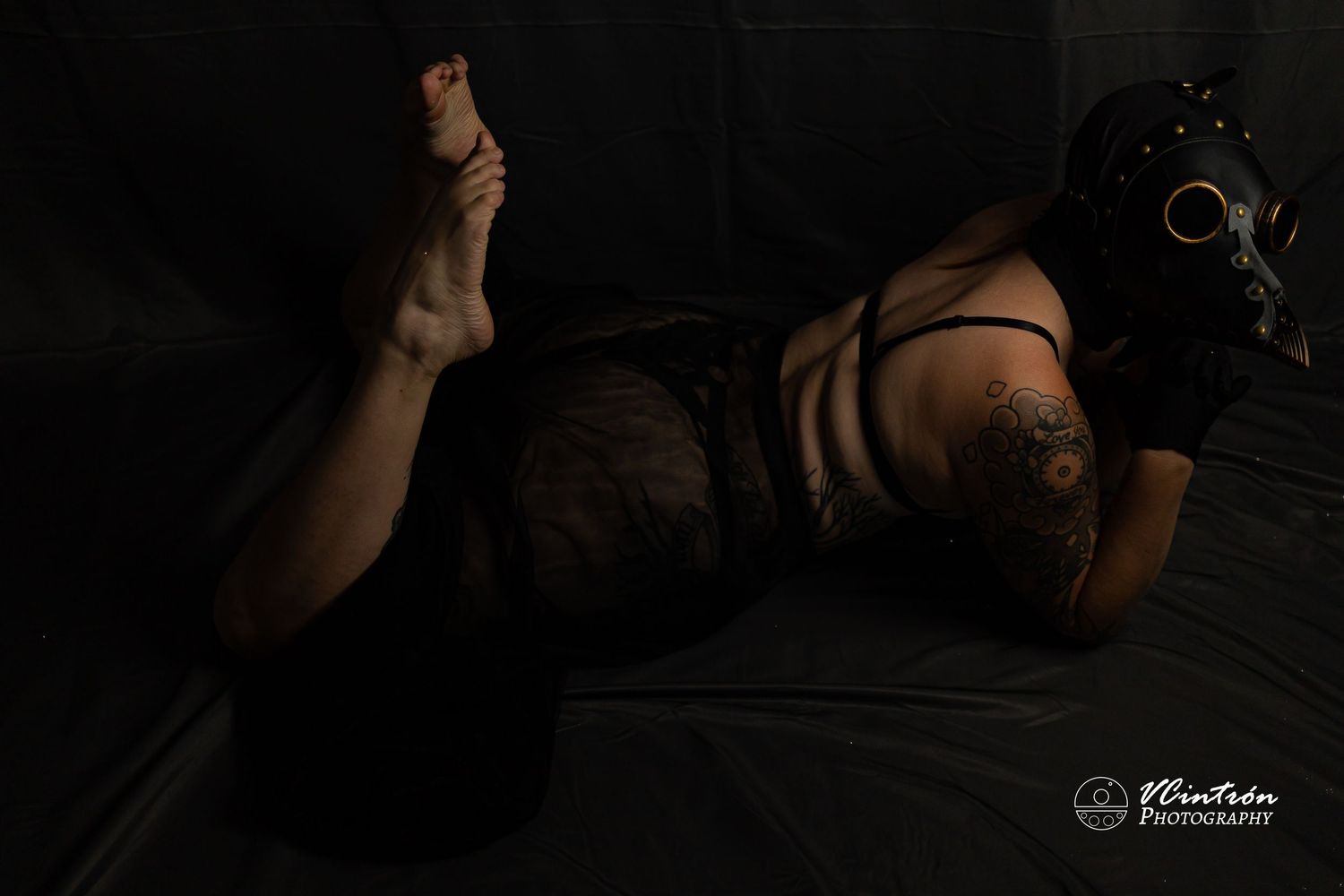 A person in black lingerie lies on a dark floor with dramatic low-key lighting creating a moody atmosphere.