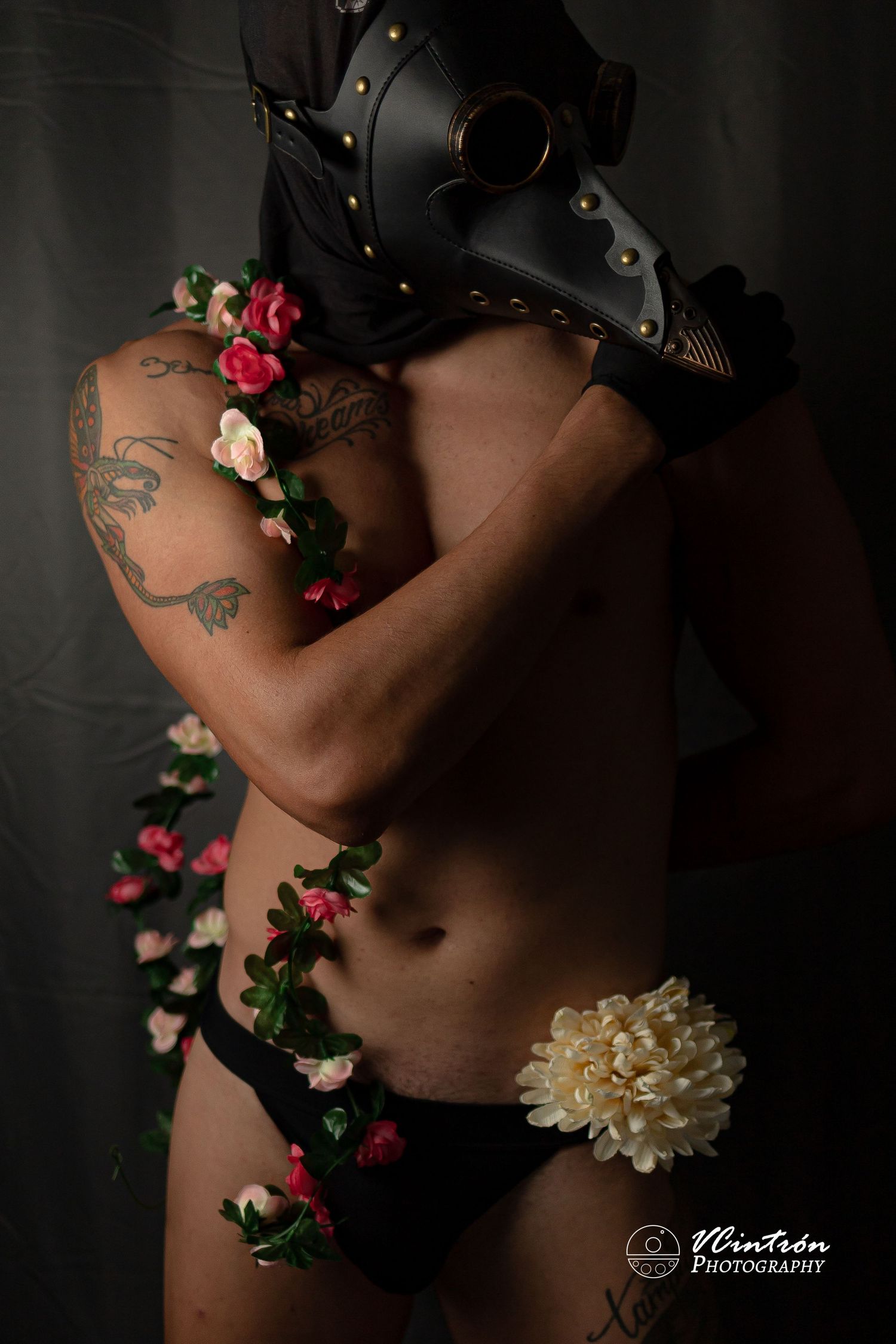 Artistic boudoir photo with black leather mask, tattoos, delicate roses and cream flower against dark background.