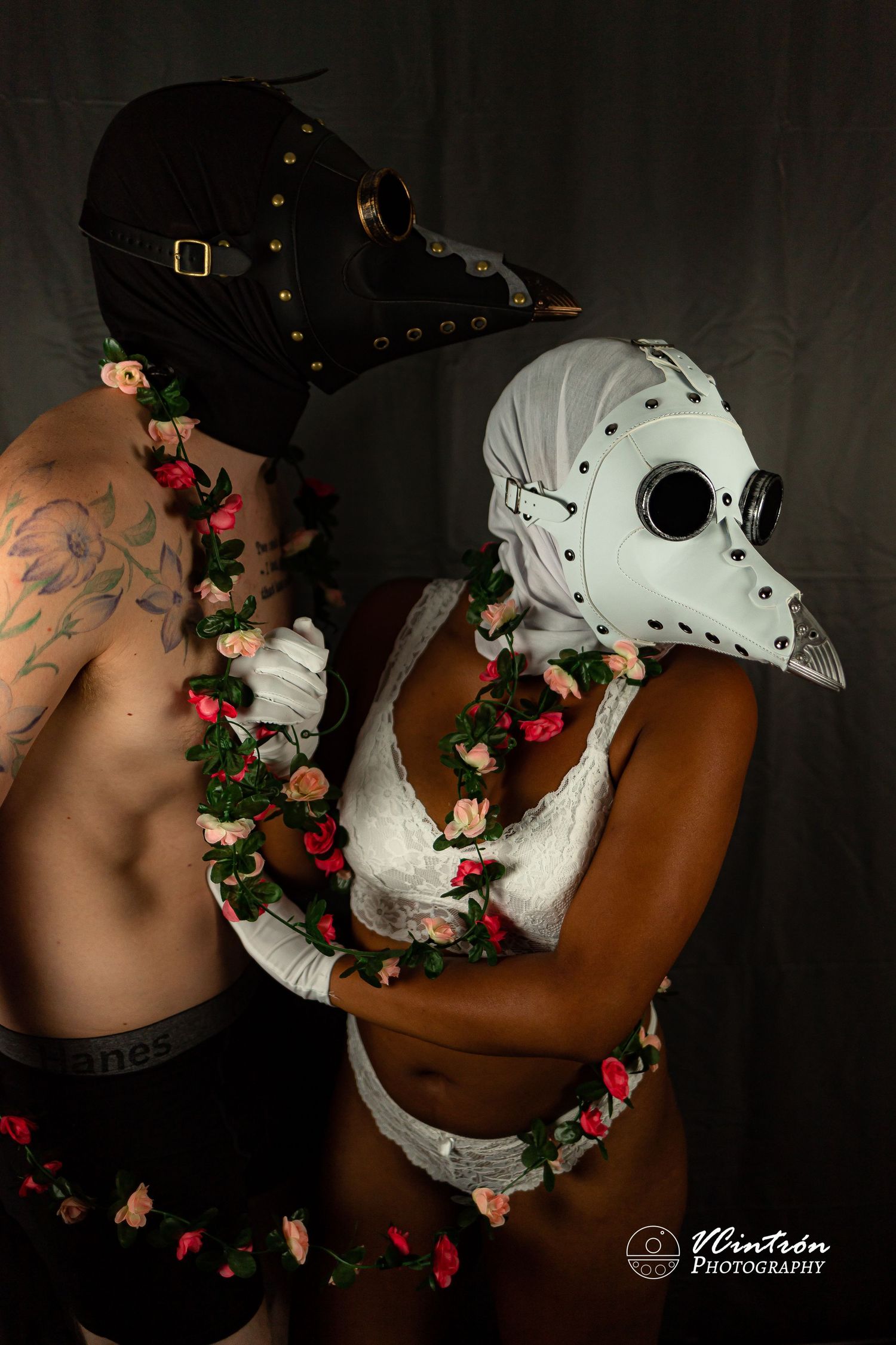 Two people wearing plague doctor and steampunk masks embrace while adorned with red rose garlands in a dark studio setting.