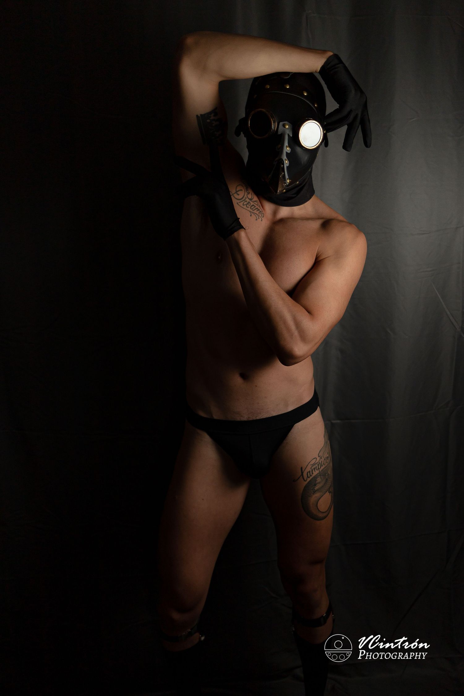 Dark and moody artistic portrait of figure in black underwear holding camera with flash against shadowy wall.