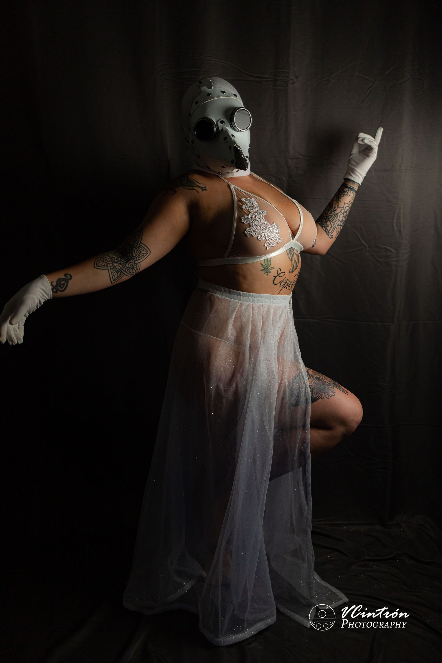Figure in white sheer dress and skull mask poses dramatically in moody dark studio lighting.