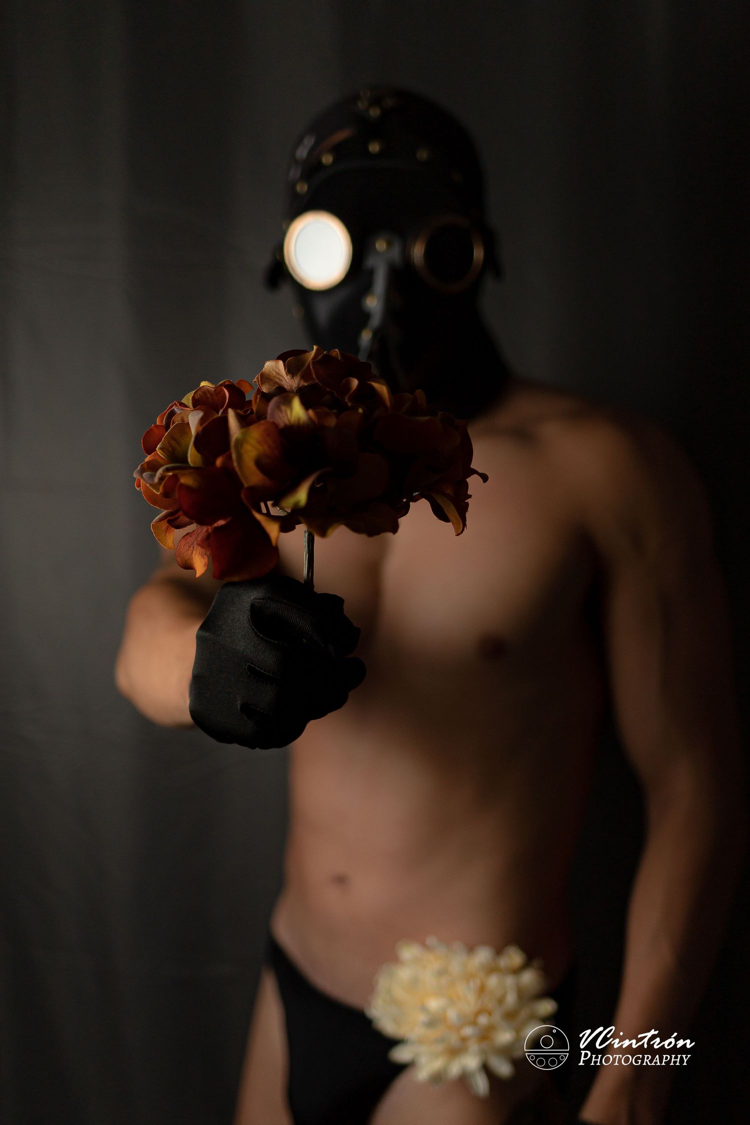 Dark artistic portrait with gas mask holding withered roses against moody backdrop and dramatic lighting.