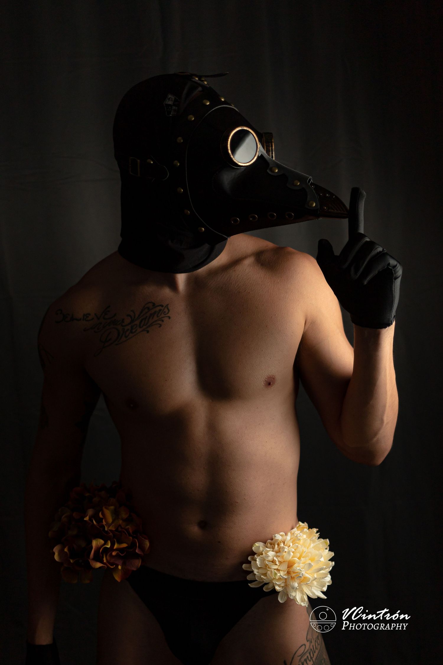 Artistic portrait featuring a person in black plague doctor mask and gloves with dramatic lighting and a white flower accent.