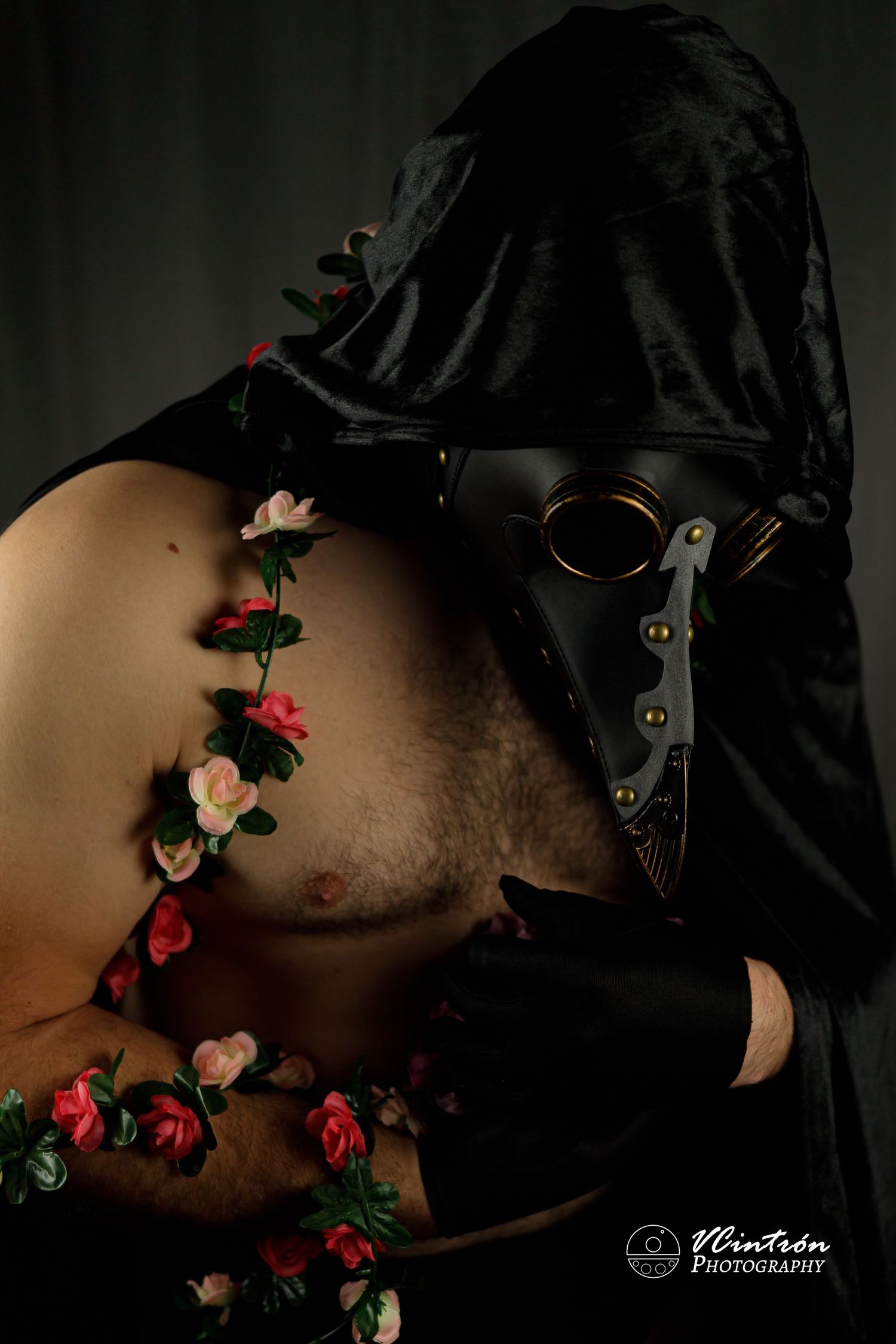 Artistic portrait showing a plague doctor mask with delicate red and pink floral vines against dark fabric.