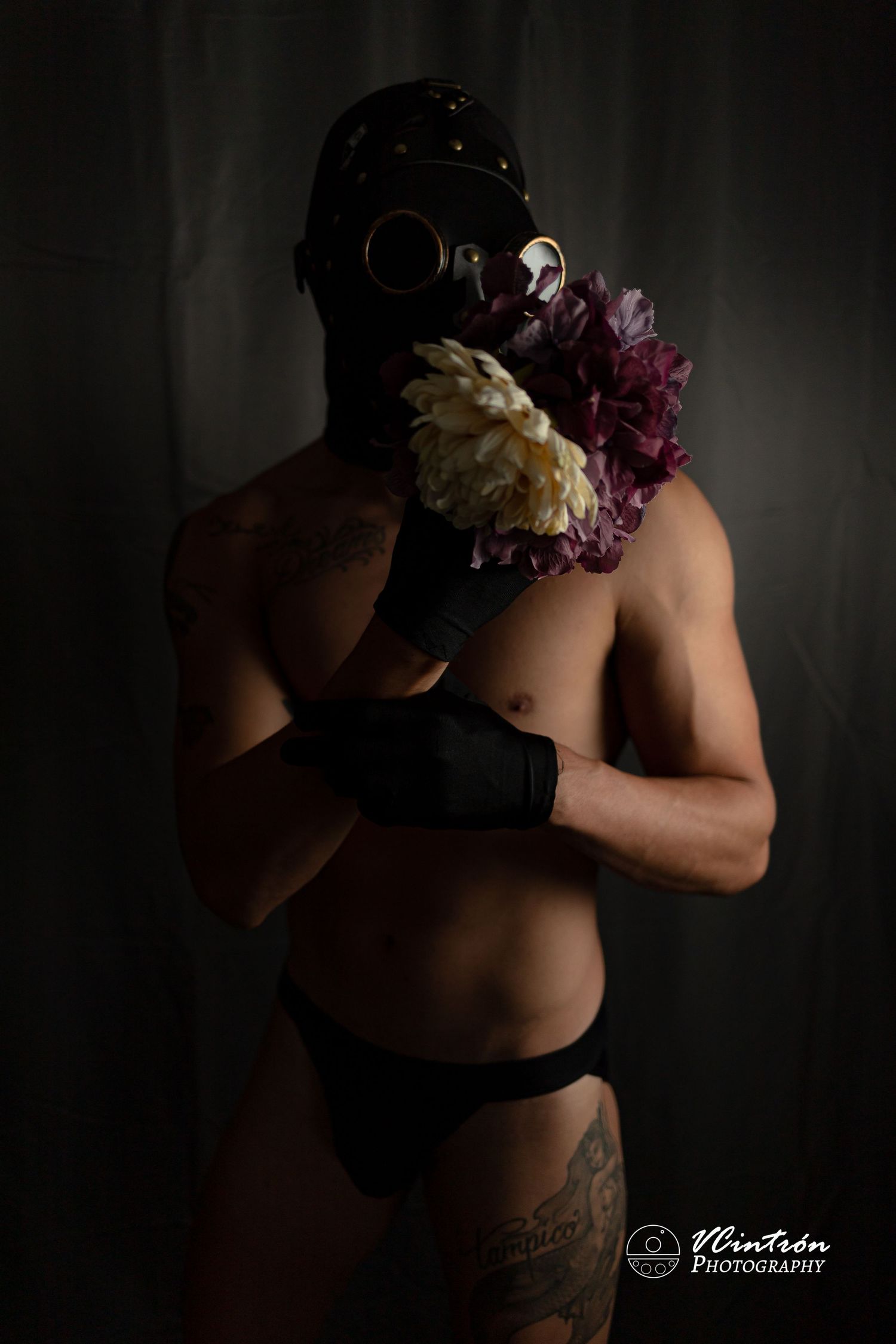 Dark artistic portrait showing figure in gas mask and black attire holding purple and cream flowers against dark backdrop.