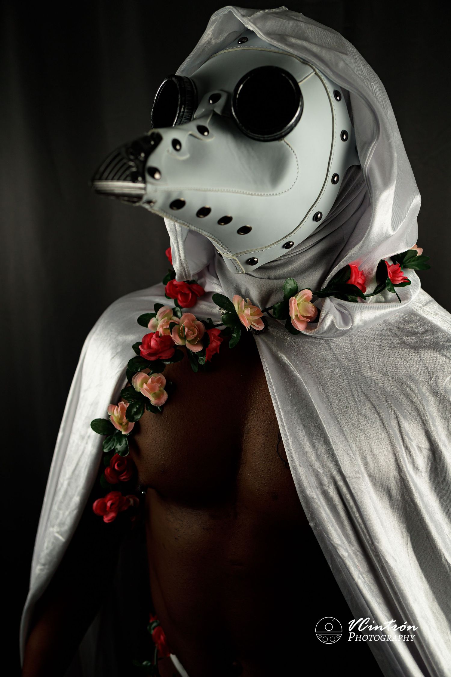 Artistic portrait of a figure wearing a white plague doctor mask adorned with floral decorations against dark background.