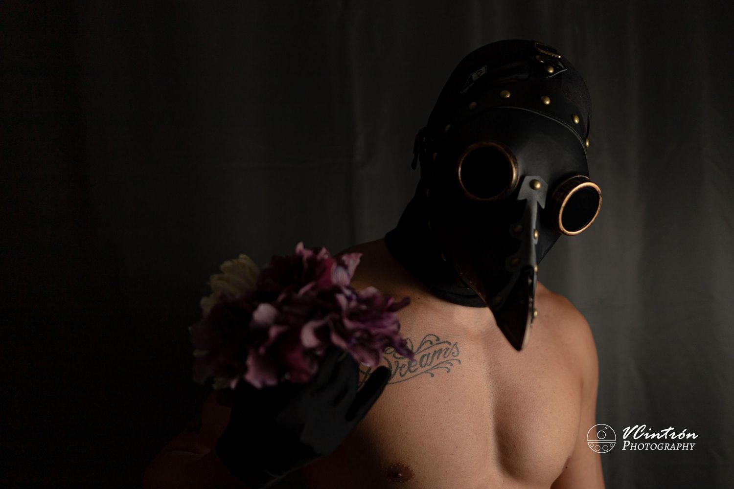 A dark artistic photo showing a plague doctor mask and shoulder tattoo with purple flowers against a black background.