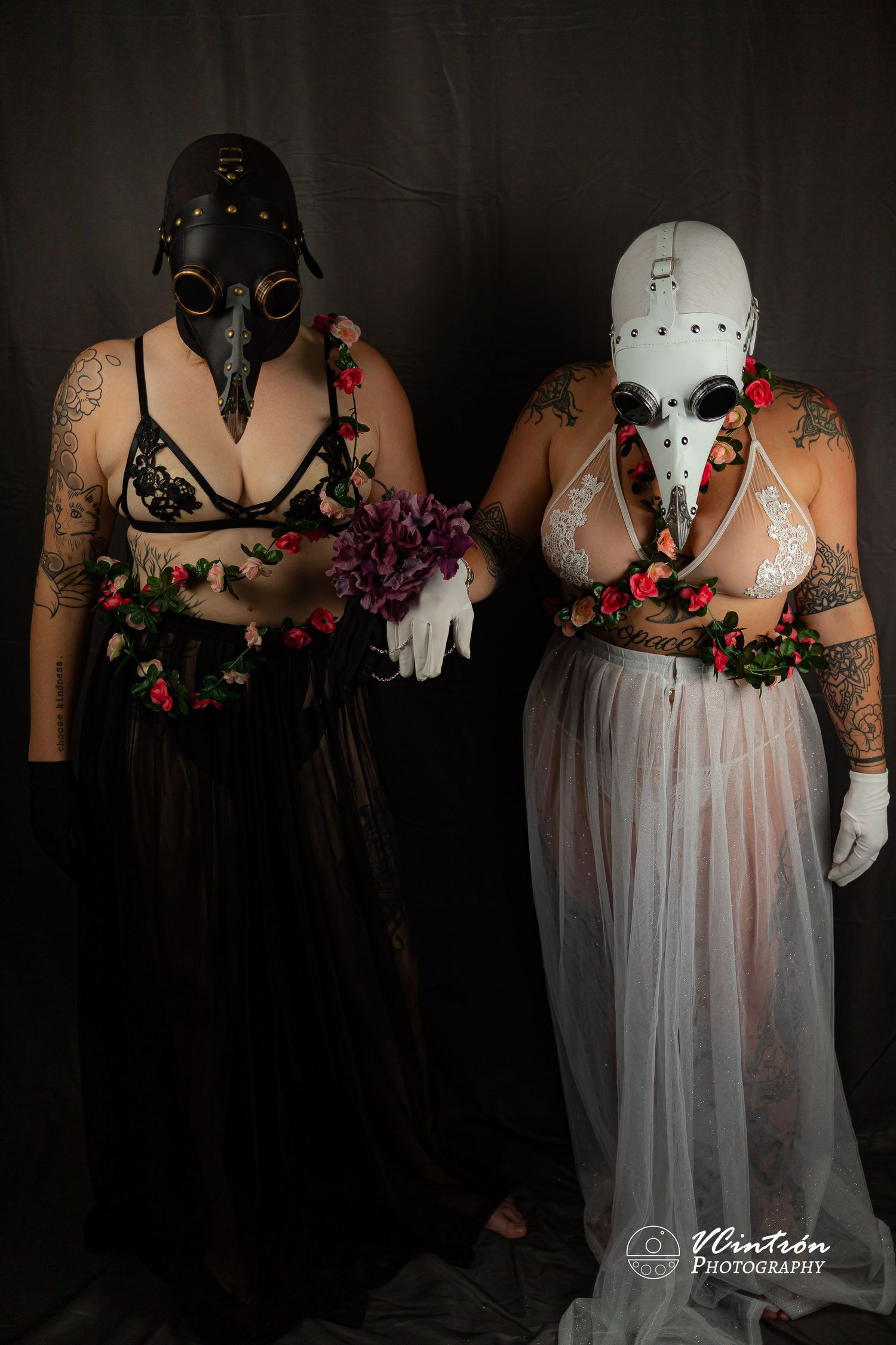 Dark artistic portrait of two figures in masks and flowing garments adorned with floral elements against a moody background.