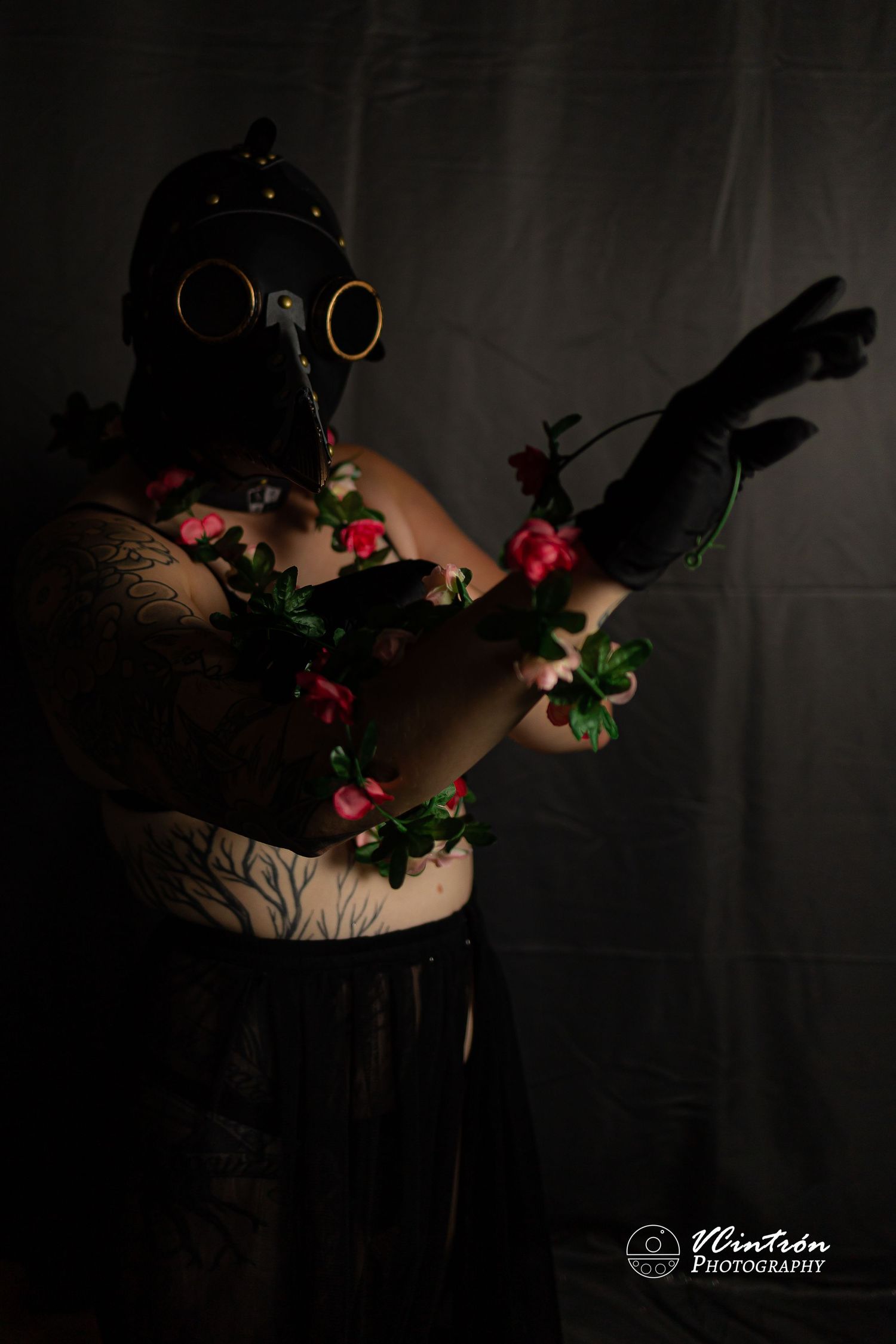 Artistic dark portrait with gas mask, draped in delicate pink roses against shadowy background with dramatic lighting.