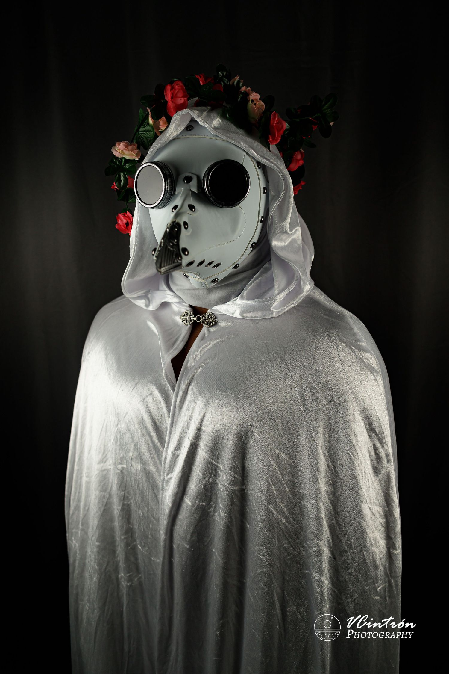 Haunting figure in white cloak wearing plague doctor mask and red flower crown against dark background.