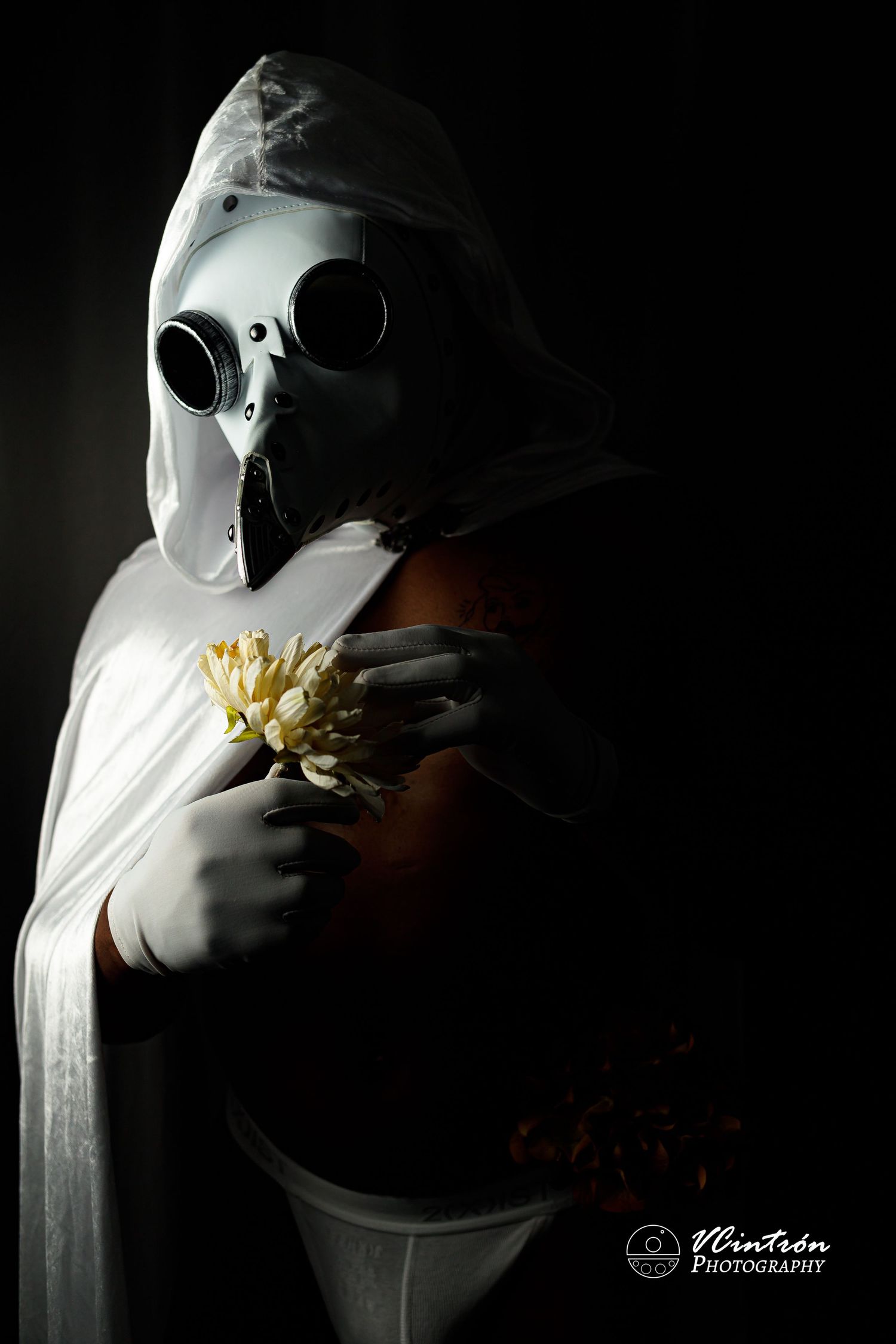 Haunting figure in plague doctor mask and white cloak holds yellow chrysanthemum flower against dark background.