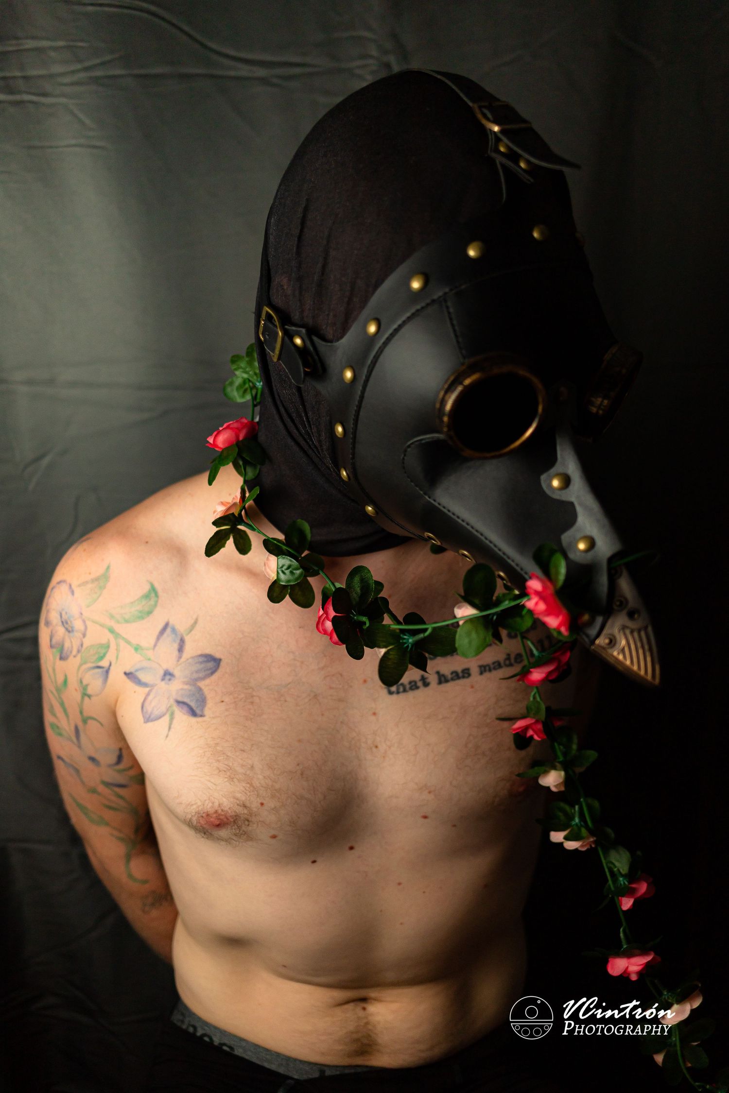 Artistic portrait showing torso with floral tattoo wearing black plague doctor mask adorned with roses and vines against dark backdrop.