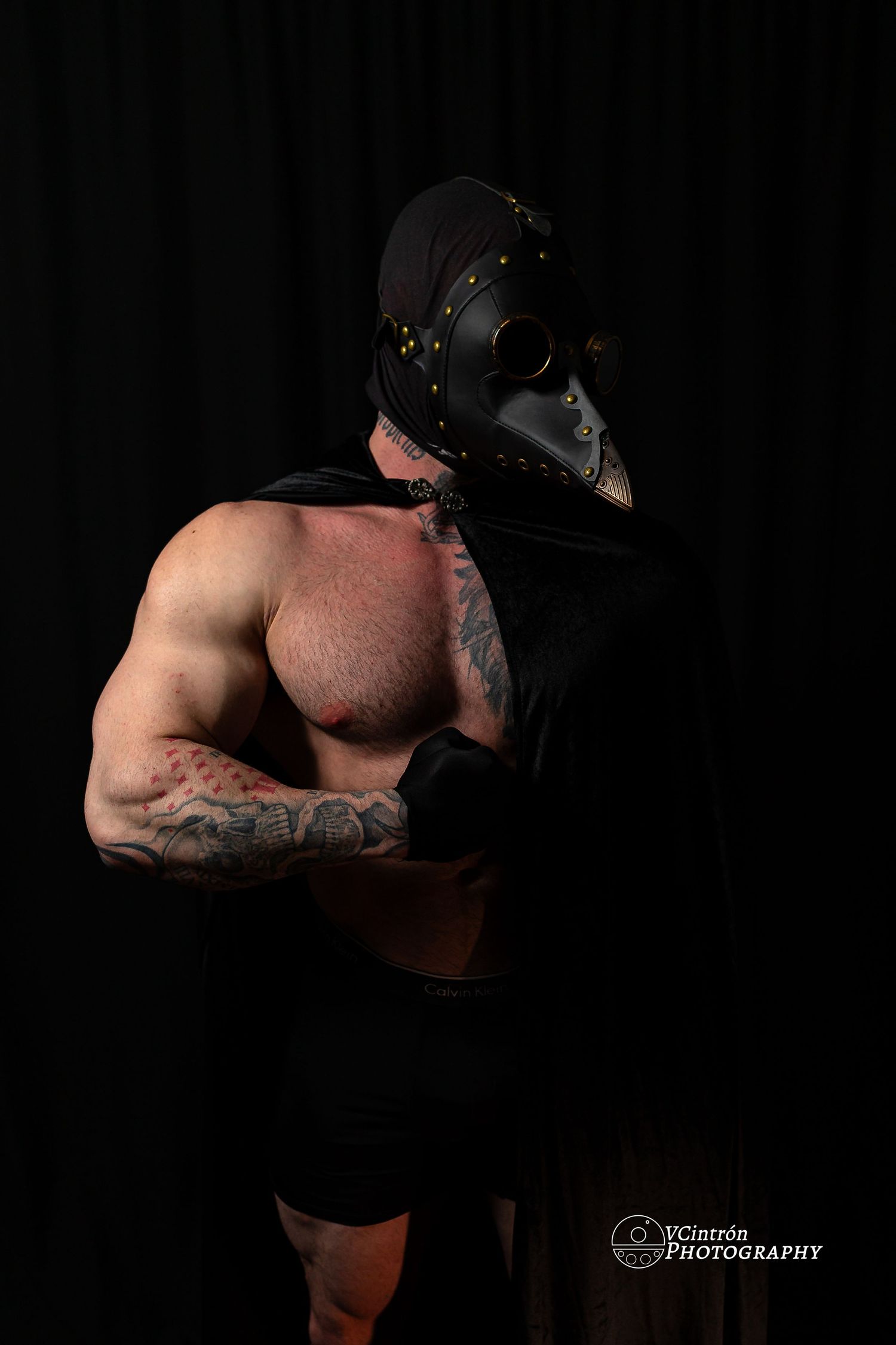 Muscular shirtless figure wearing black leather gladiator mask poses dramatically against dark background in studio lighting.